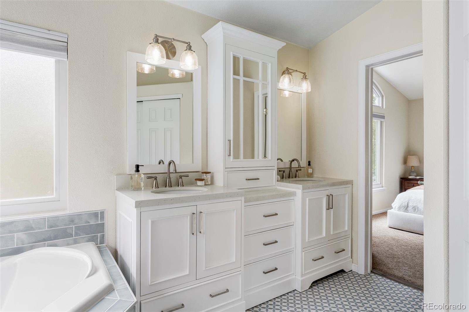 Bathroom with double white vanities, a built-in mirrored cabinet, patterned tile floor, and tiled bathtub surround.