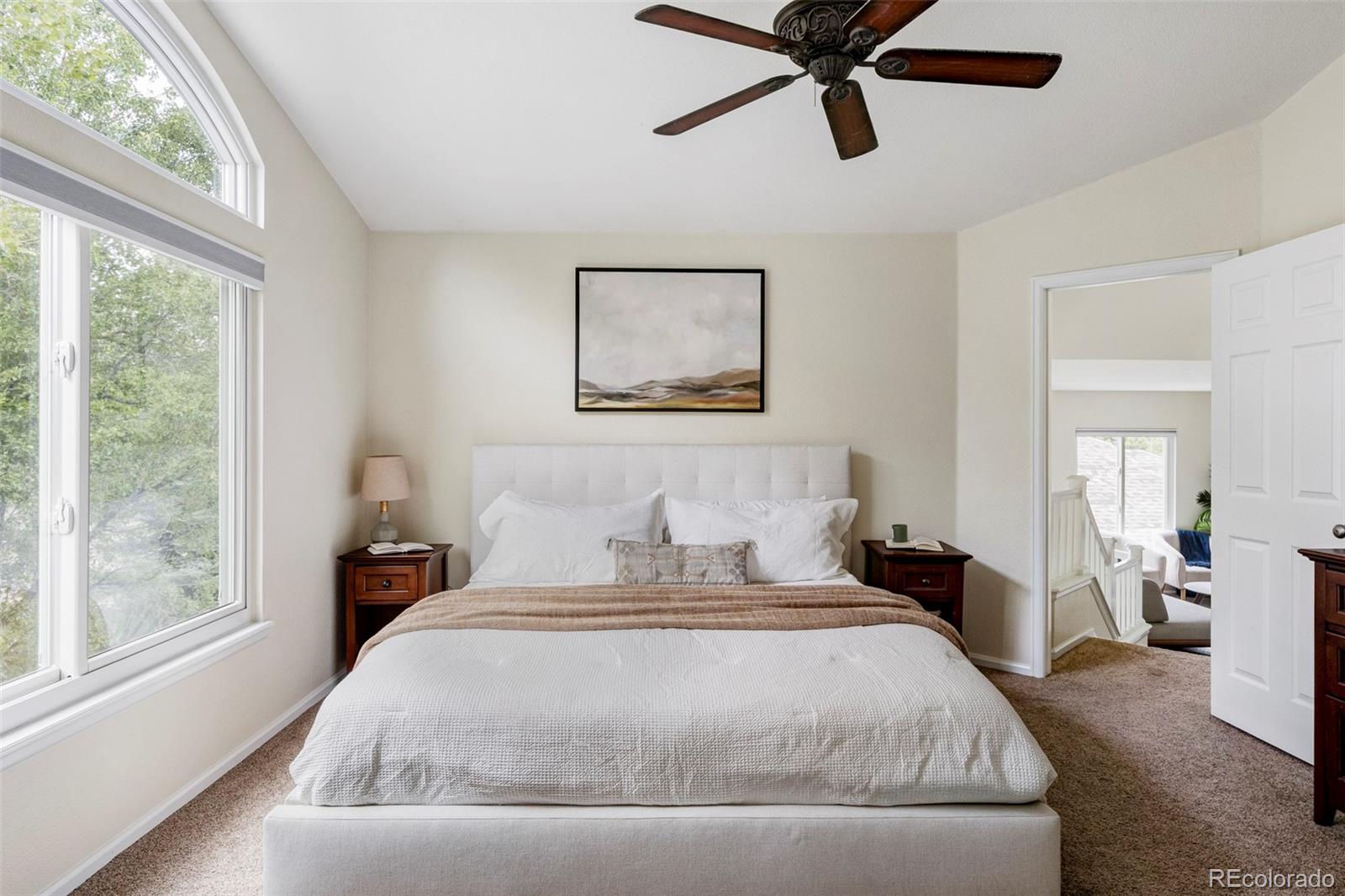 Furnished bedroom with a large white bed, tall arched windows, and a dark wood ceiling fan.