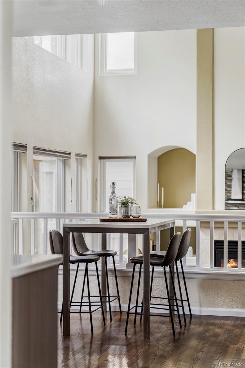 Dining area with a tall table and stools beside large windows, overlooking a living room fireplace.