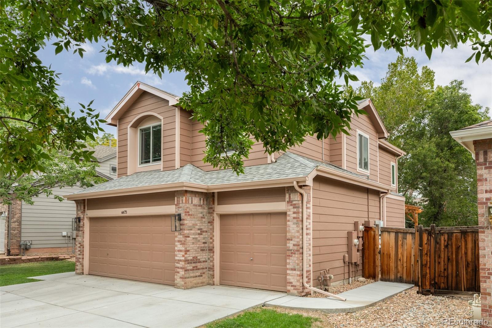 Two-story tan house with brick accents, two garage doors, and leafy trees overhead.