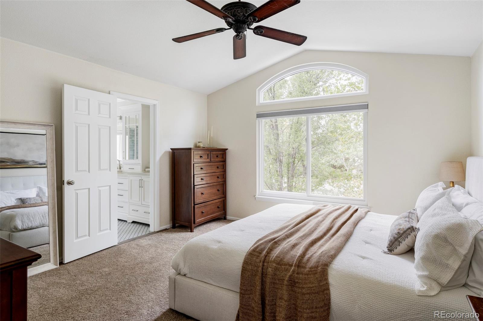 Bedroom with a large arched window, dark wood chest, and open bathroom doorway.