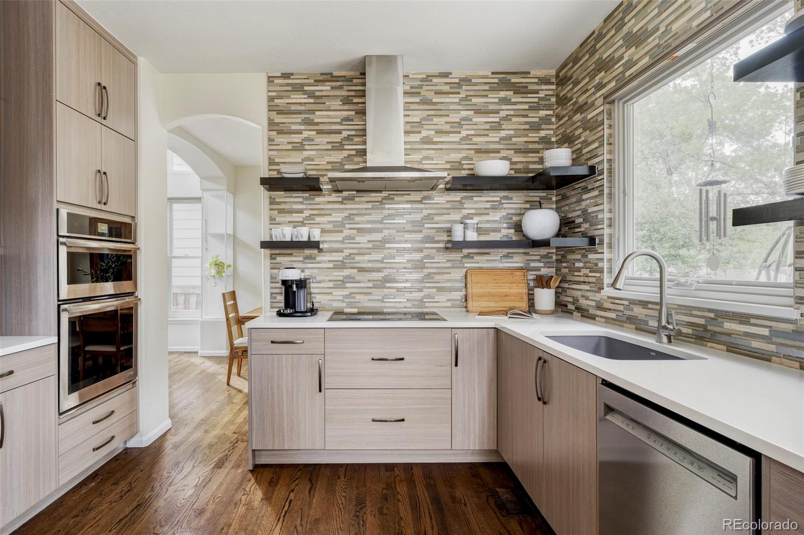 Kitchen with multicolored tile backsplash, light wood cabinets, stainless appliances, and a large window over the sink.