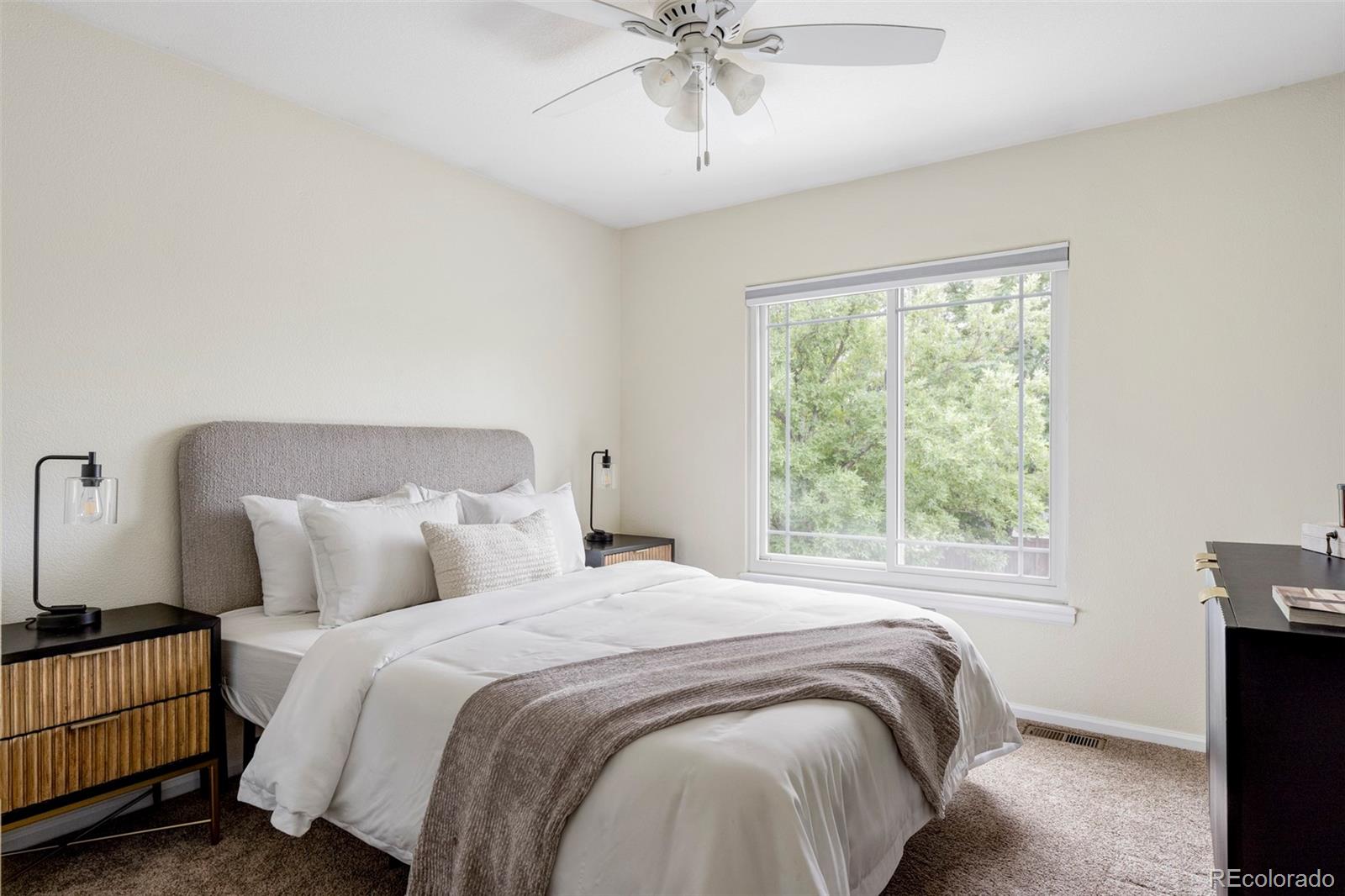 Staged bedroom with a gray upholstered headboard and a large window overlooking leafy trees.