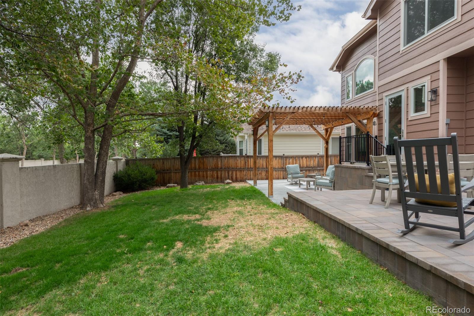 Backyard patio with a wooden pergola, outdoor seating, mature trees, and a green lawn.