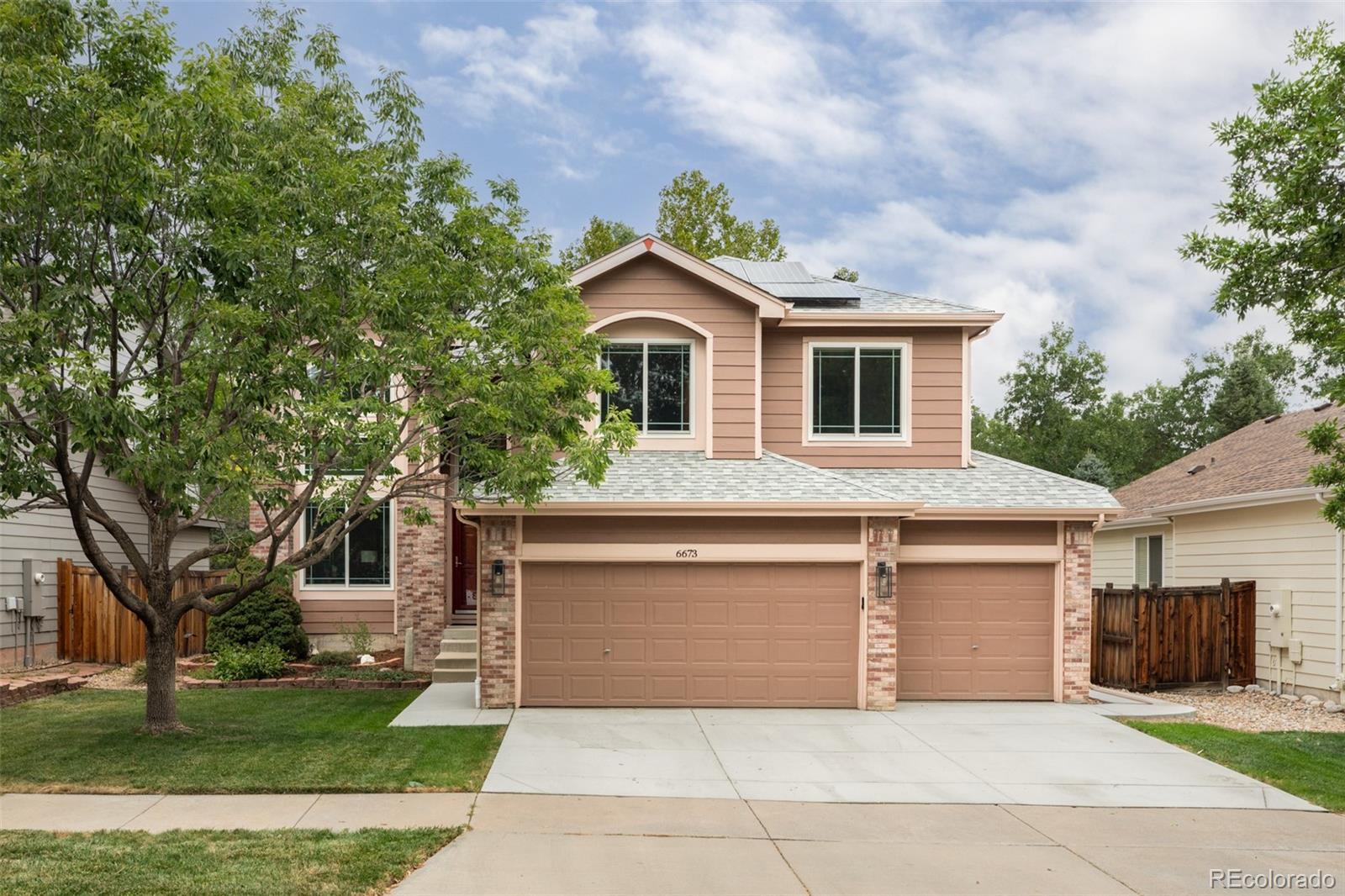 Two-story tan and brick house with solar panels, a two-car garage, and mature trees in front.
