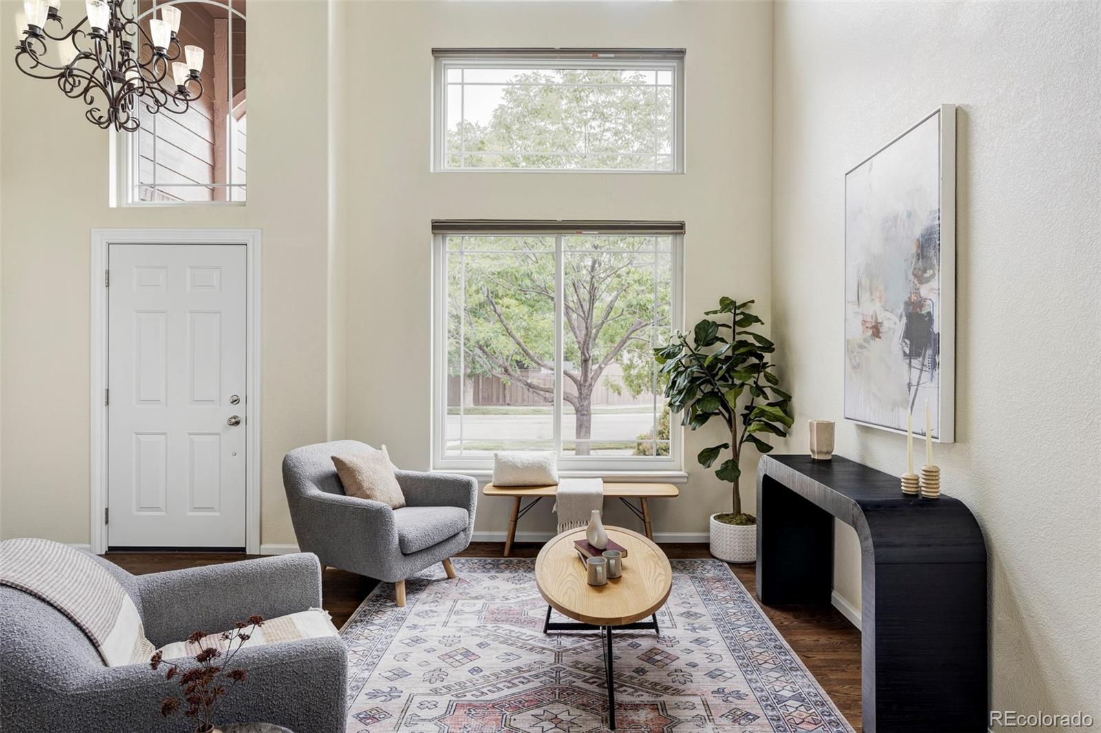 Staged living room with tall windows, gray armchairs, a patterned rug, and a dark fireplace console.