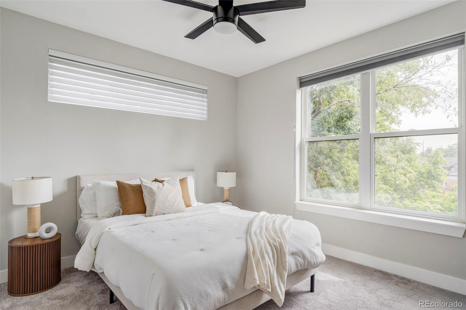 Staged bedroom with a white bed, dark ceiling fan, and large windows overlooking leafy trees.