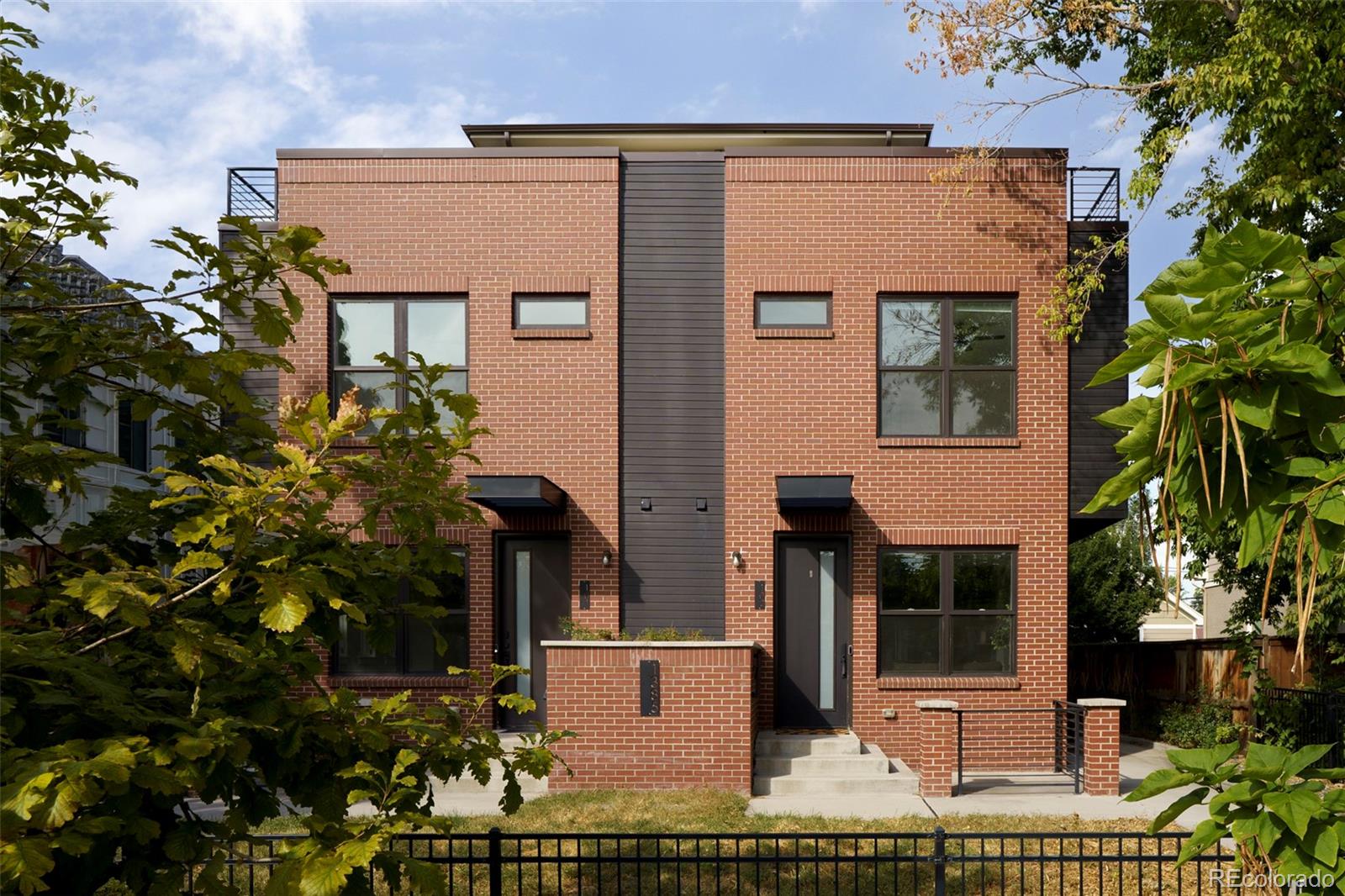 Modern red-brick townhouse with dark siding, upper balconies, and leafy trees.