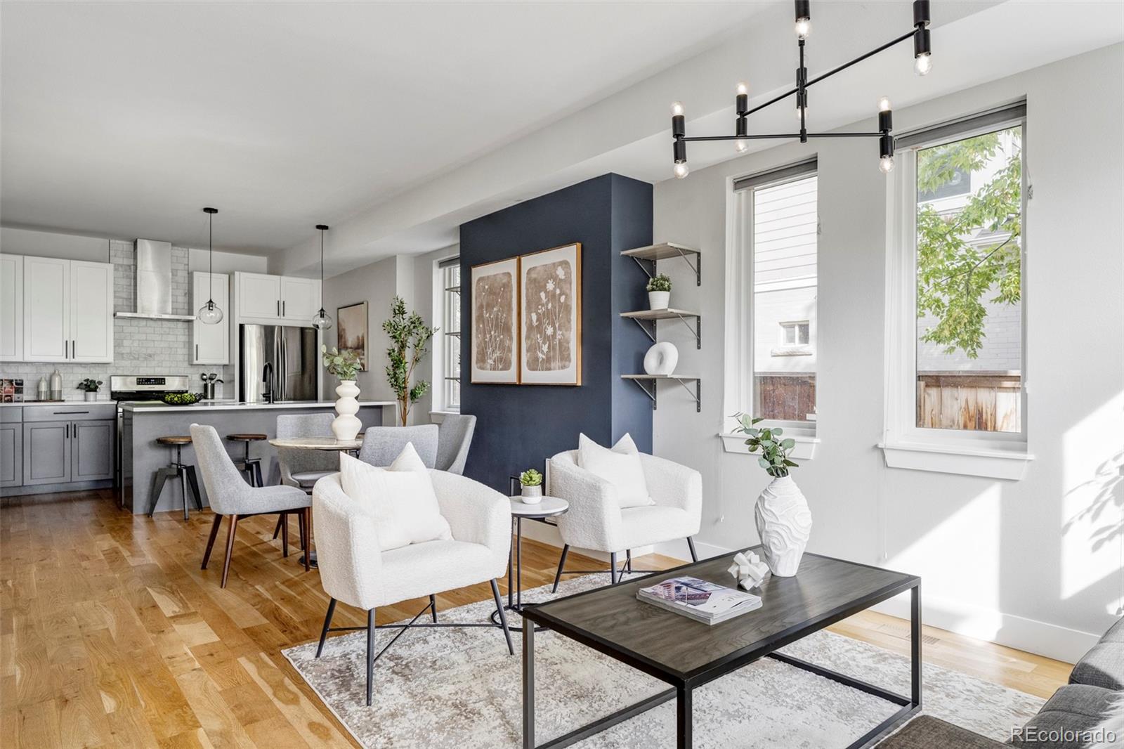Open-plan living room with white chairs, hardwood floors, navy accent wall, and large windows.