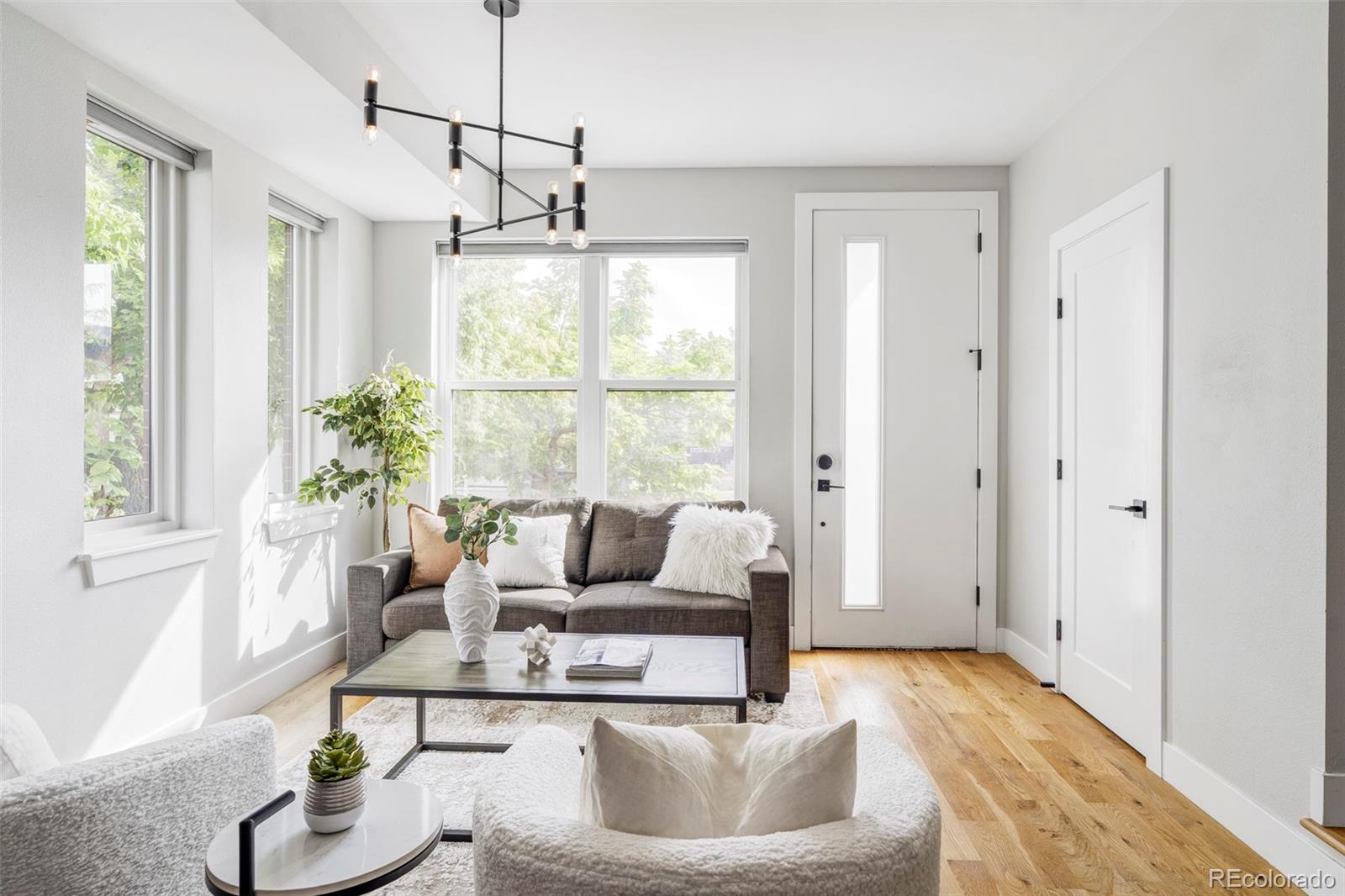 Bright staged living room with large windows, a gray sofa, white chairs, and light wood floors.