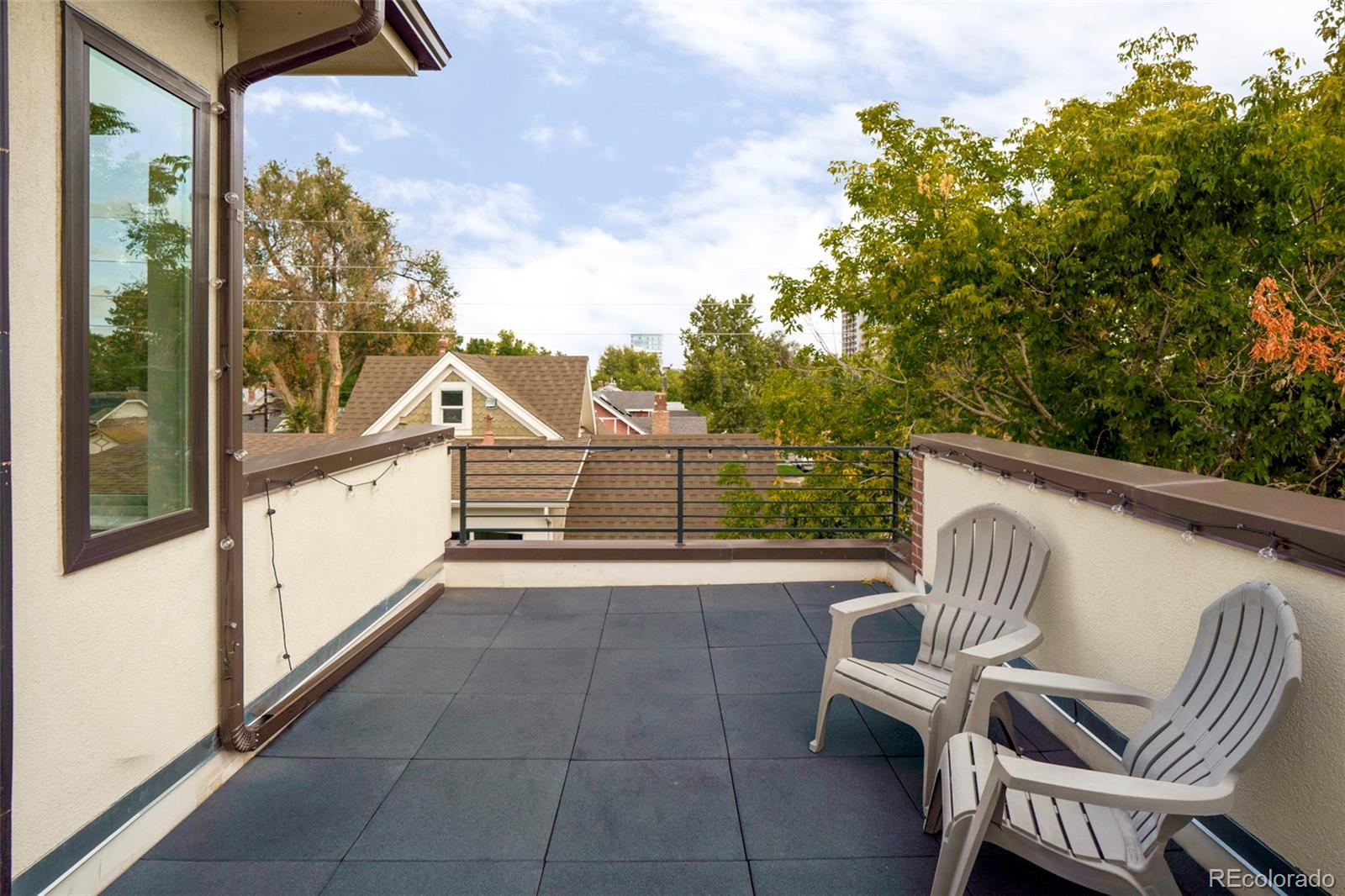 Rooftop terrace with dark pavers, white lounge chairs, string lights, and leafy trees beyond the railing.
