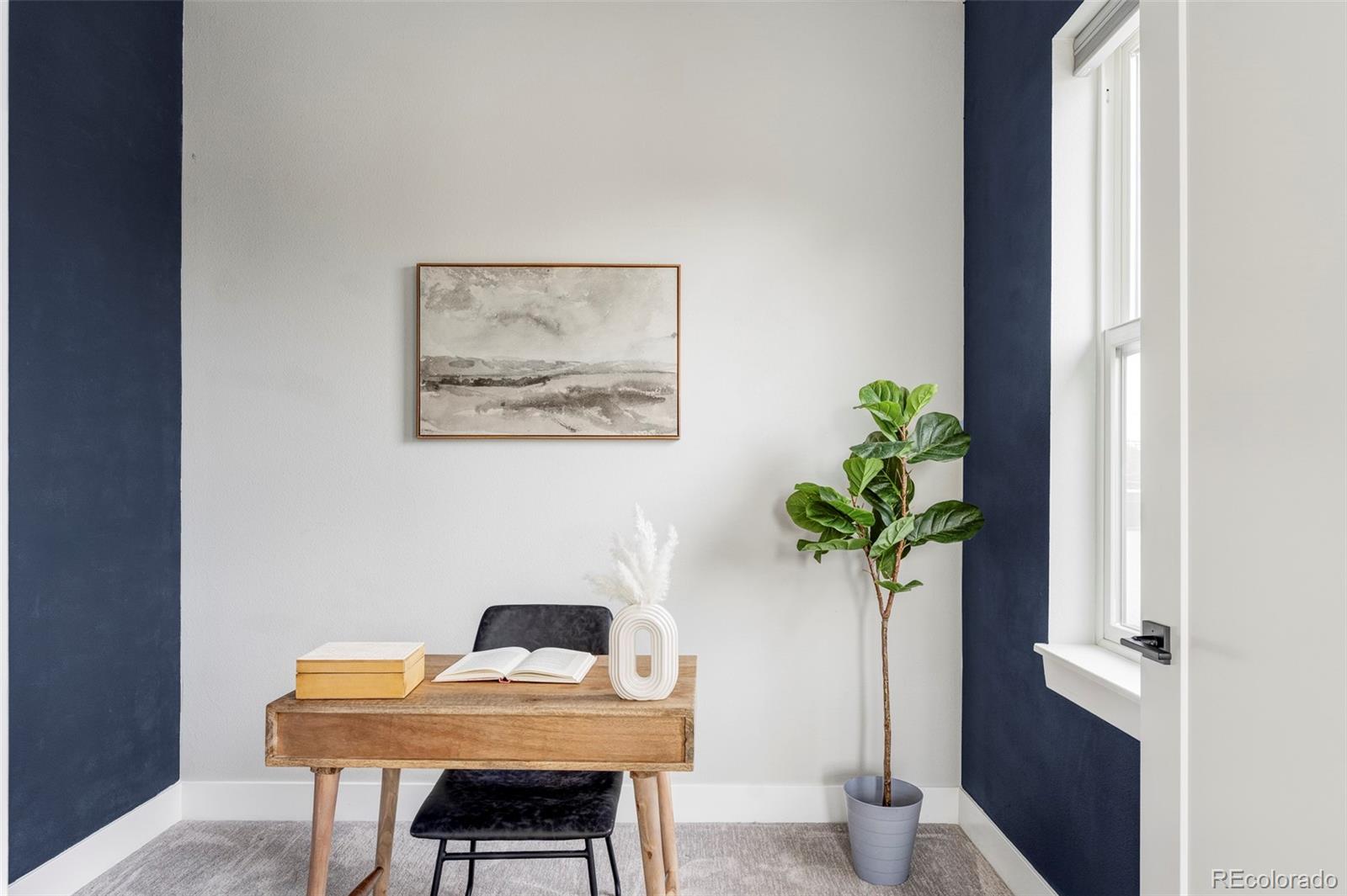 Home office with a wooden desk, navy accent walls, framed art, and a tall window.