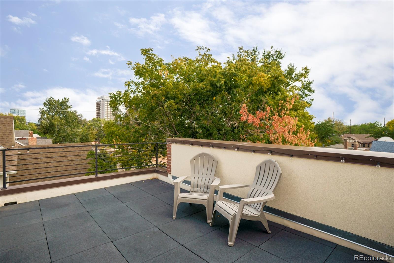 Rooftop terrace with two white chairs, dark pavers, and autumn trees beyond the railing.