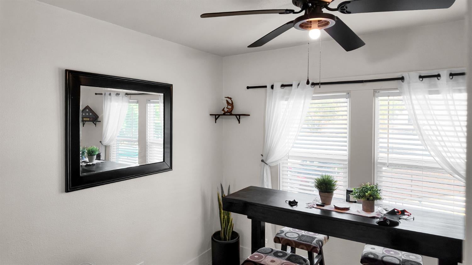 Dining area with a dark table, patterned stools, bright curtained windows, and a ceiling fan.