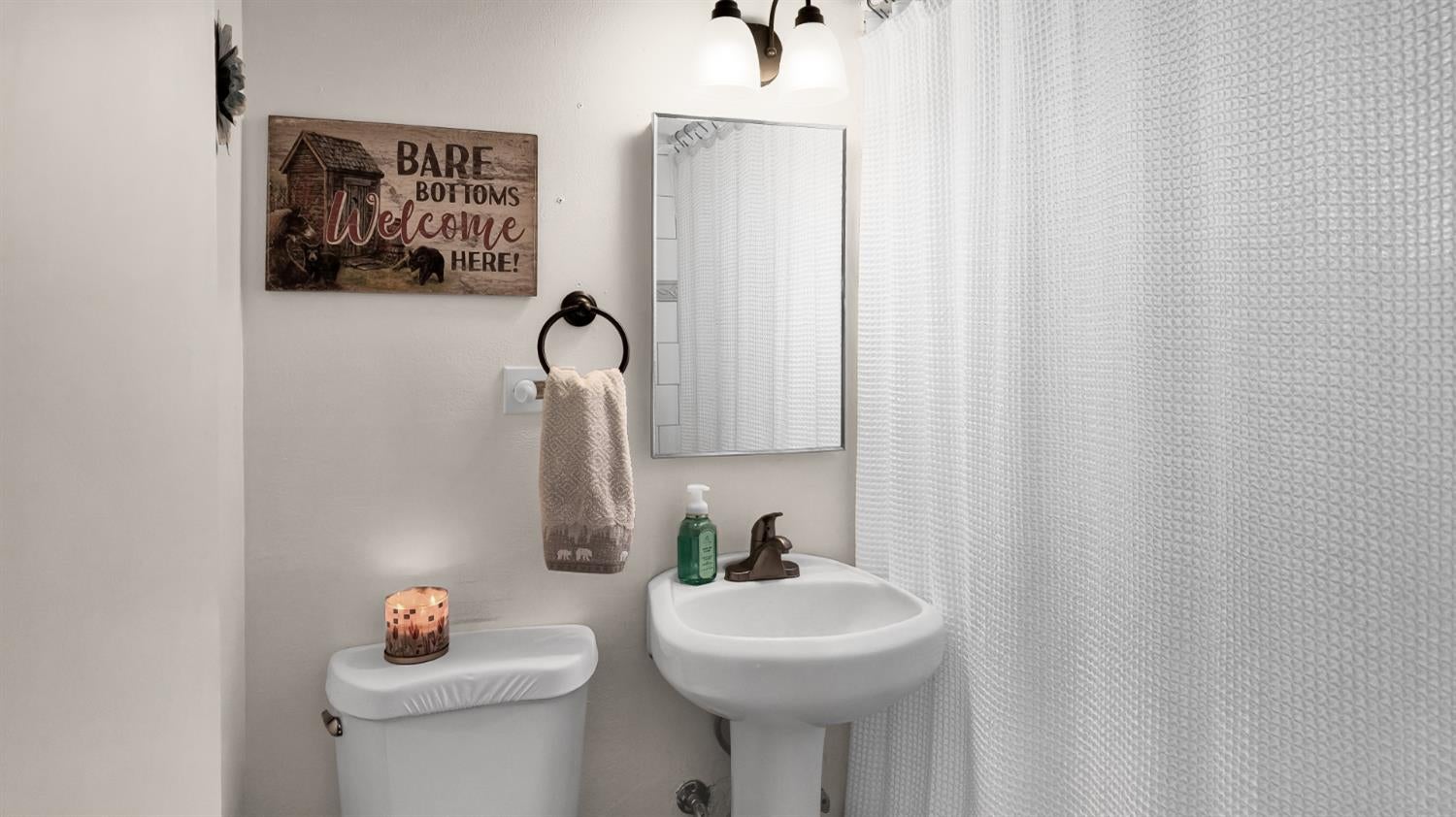 Bathroom with a white pedestal sink, toilet, and textured shower curtain beside a rustic welcome sign.