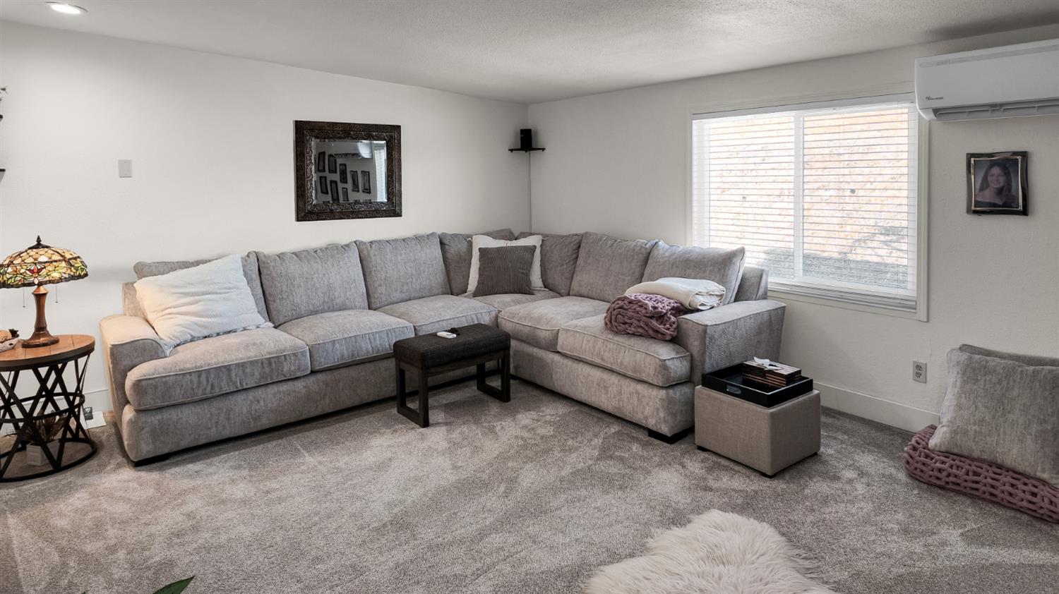 Living room with a gray sectional sofa, dark ottoman, and window with blinds.