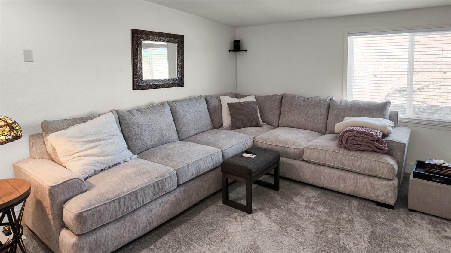 Living room with a large gray sectional sofa, dark ottoman, and wide window with blinds.