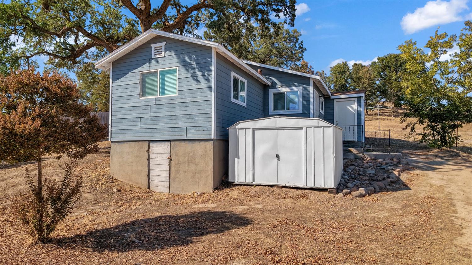 Blue-sided house on a raised foundation with a white storage shed in the dry yard.