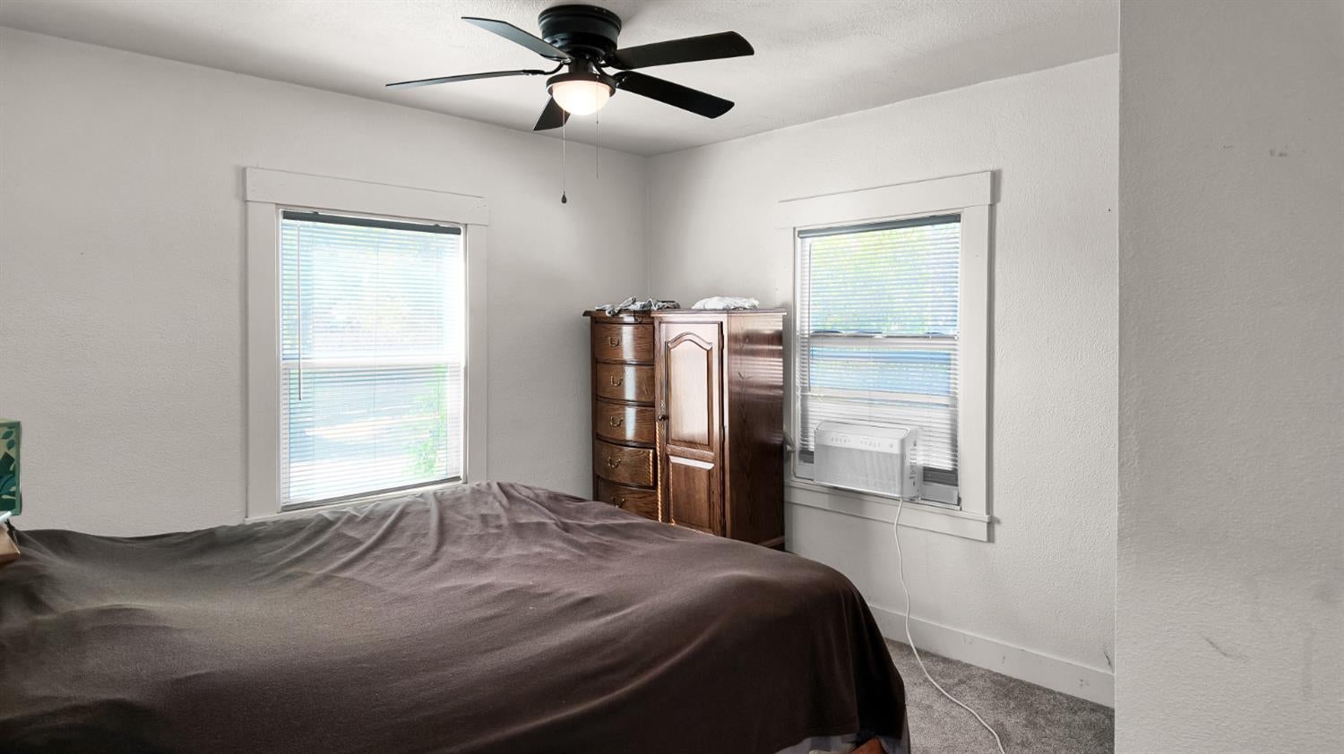 Bedroom with a dark-covered bed, wooden dresser between two windows, and ceiling fan.