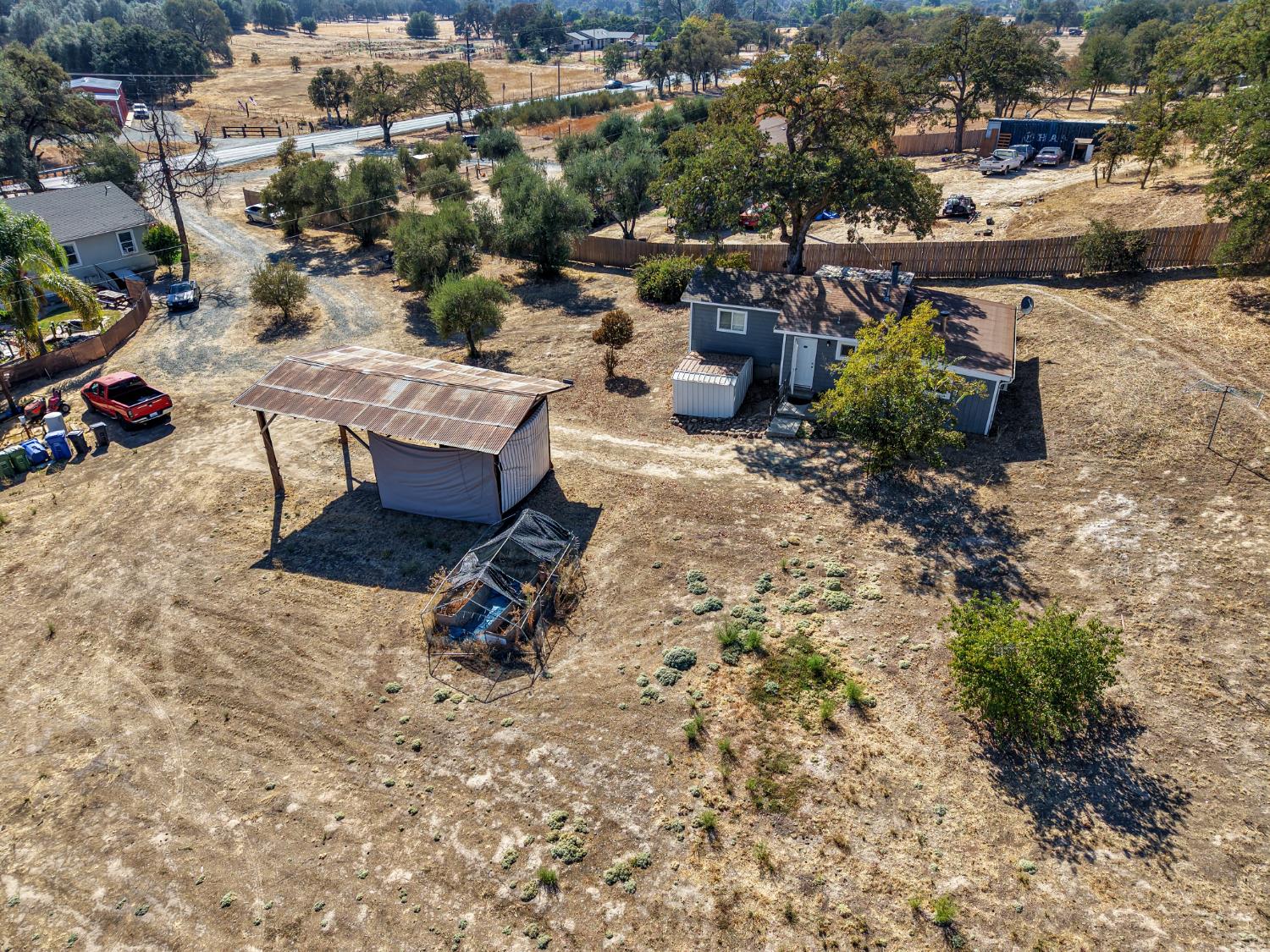 Aerial view of a rural lot with a small gray house, metal-roofed outbuilding, and dry open ground.