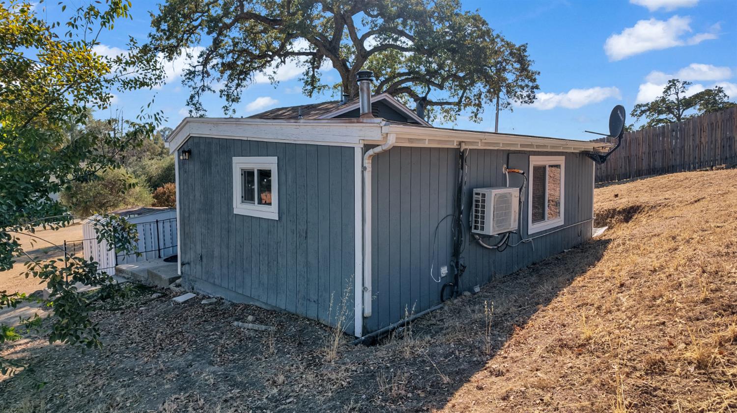 Blue-gray house with white trim on a dry slope beneath a large oak tree.