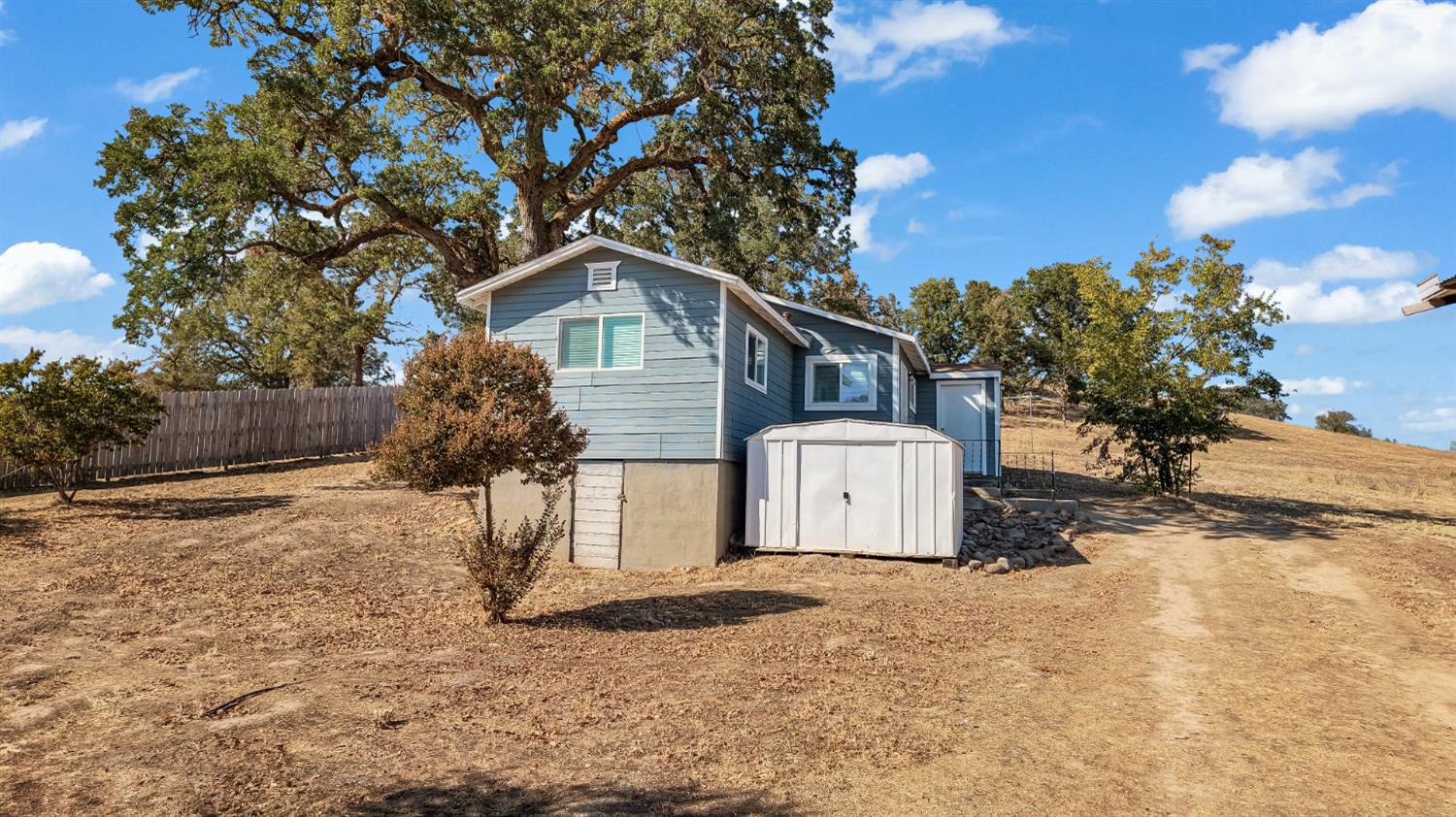 Blue-gray house with a white storage shed on a dry, oak-dotted lot.