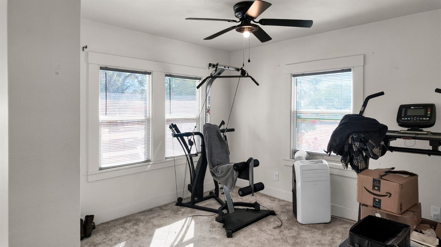 Exercise room with a multi-station gym, treadmill, and portable air conditioner by three windows.