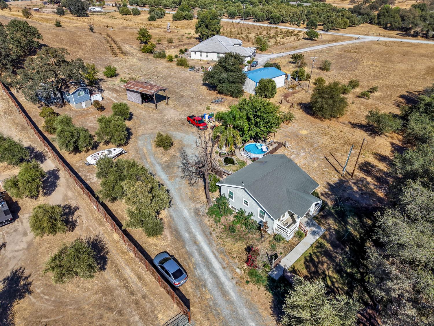 Aerial view of rural homes and outbuildings on dry acreage, with a pool and gravel driveway.