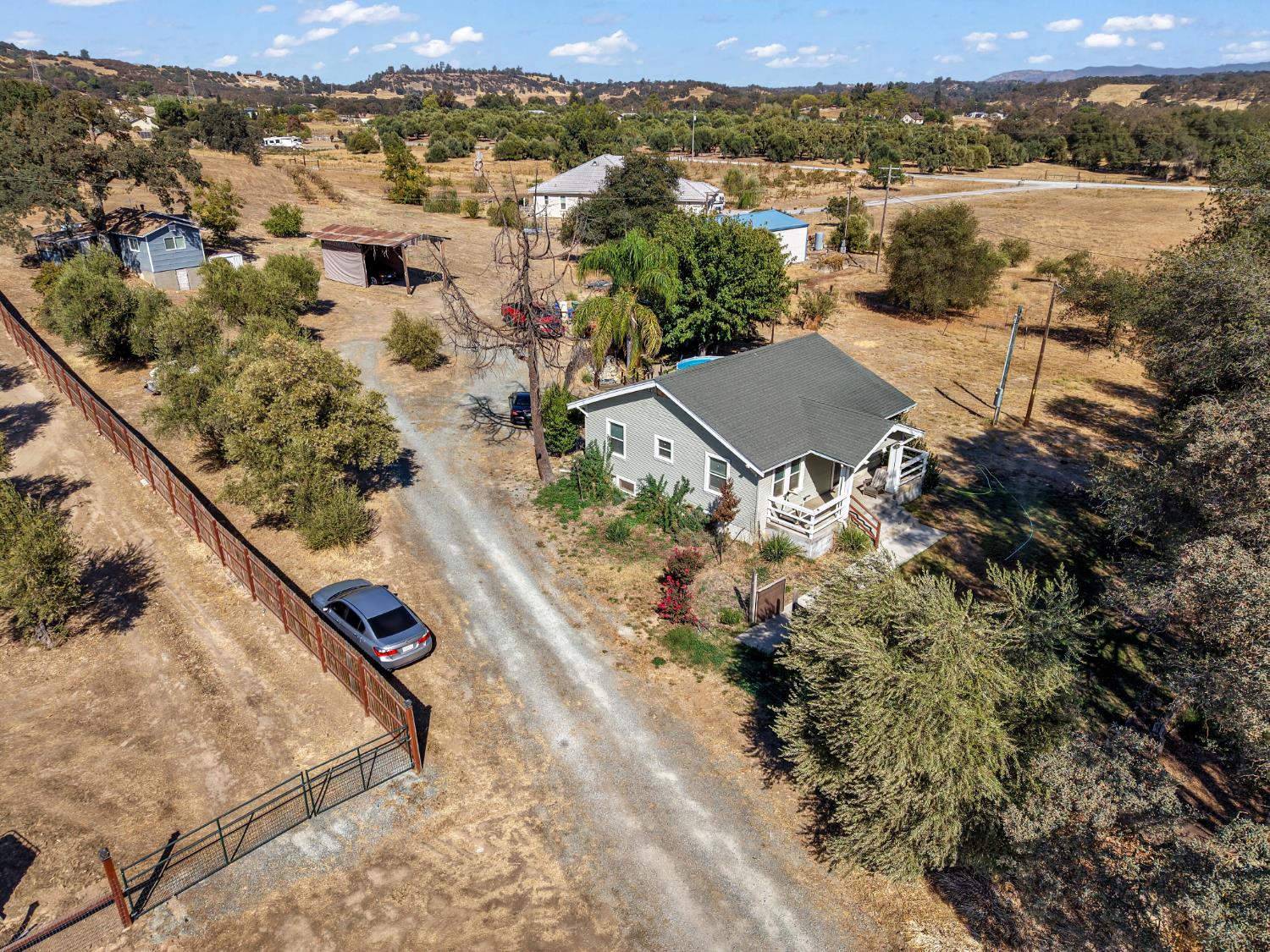 Aerial view of a gray house on a dry rural lot with outbuildings and rolling hills beyond.