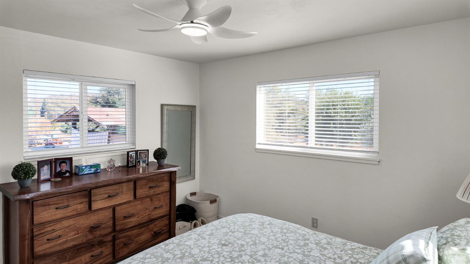 Bedroom with a patterned bed, wooden dresser, two windows with blinds, and ceiling fan.