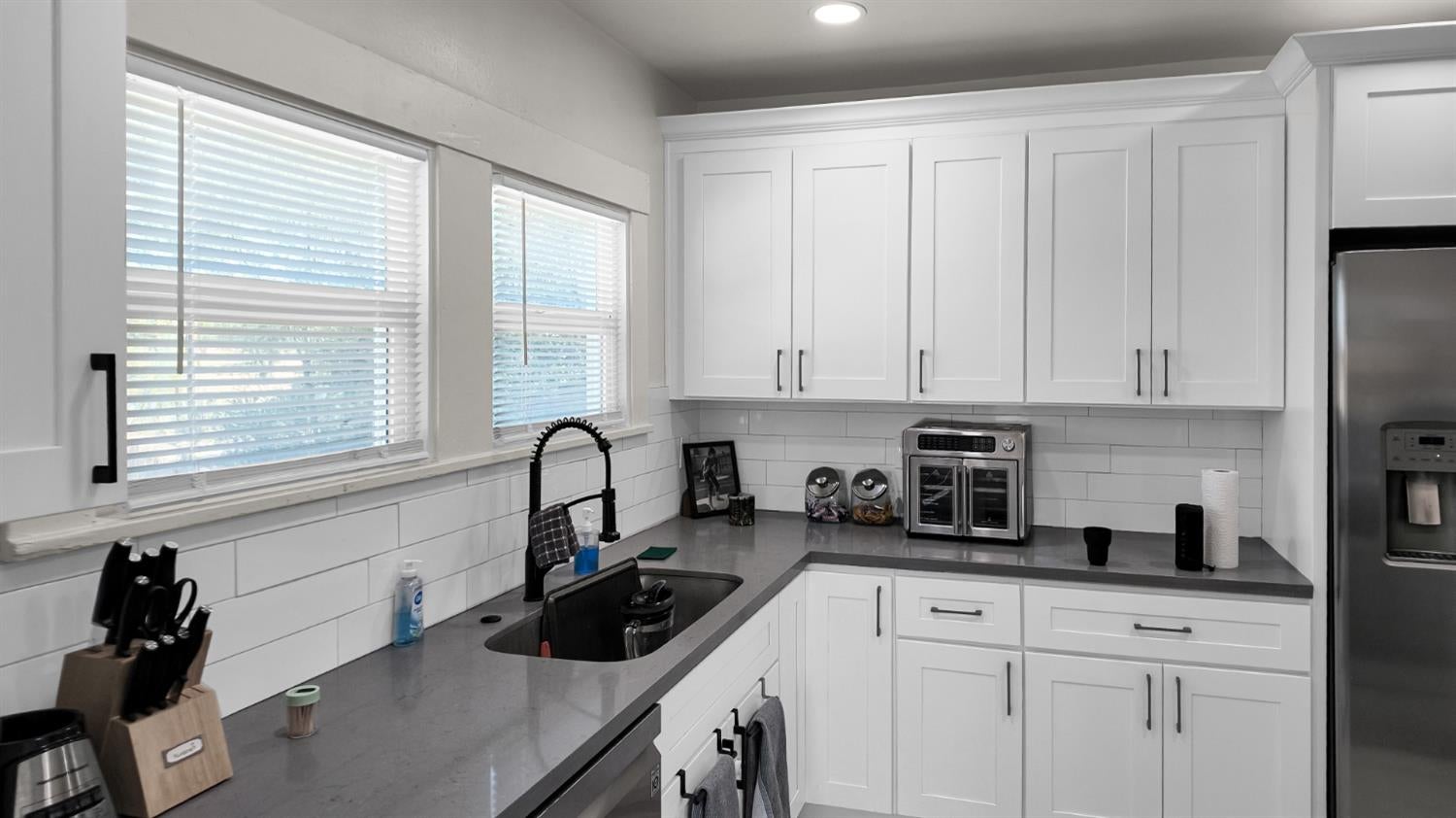 Kitchen with white shaker cabinets, gray counters, a black sink, and two windows with blinds.