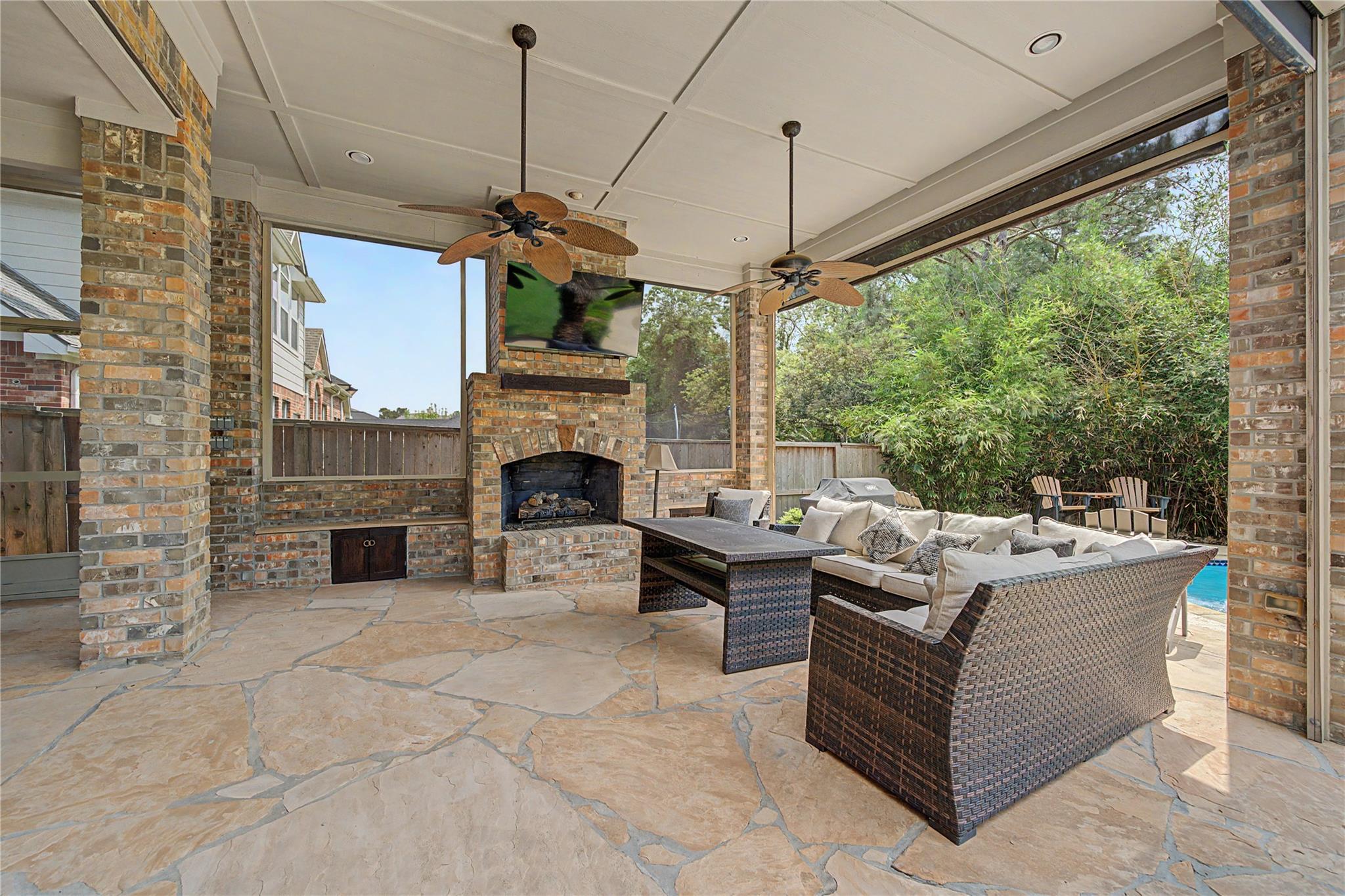 Covered patio with a brick fireplace, ceiling fans, wicker seating, and a glimpse of the pool.