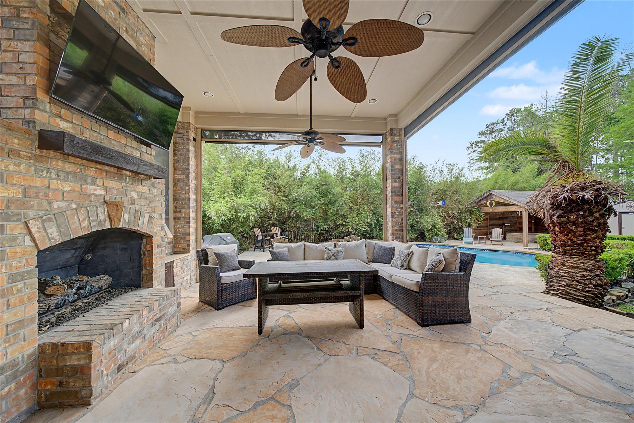 Covered patio with brick fireplace and wicker seating overlooking a swimming pool and cabana.