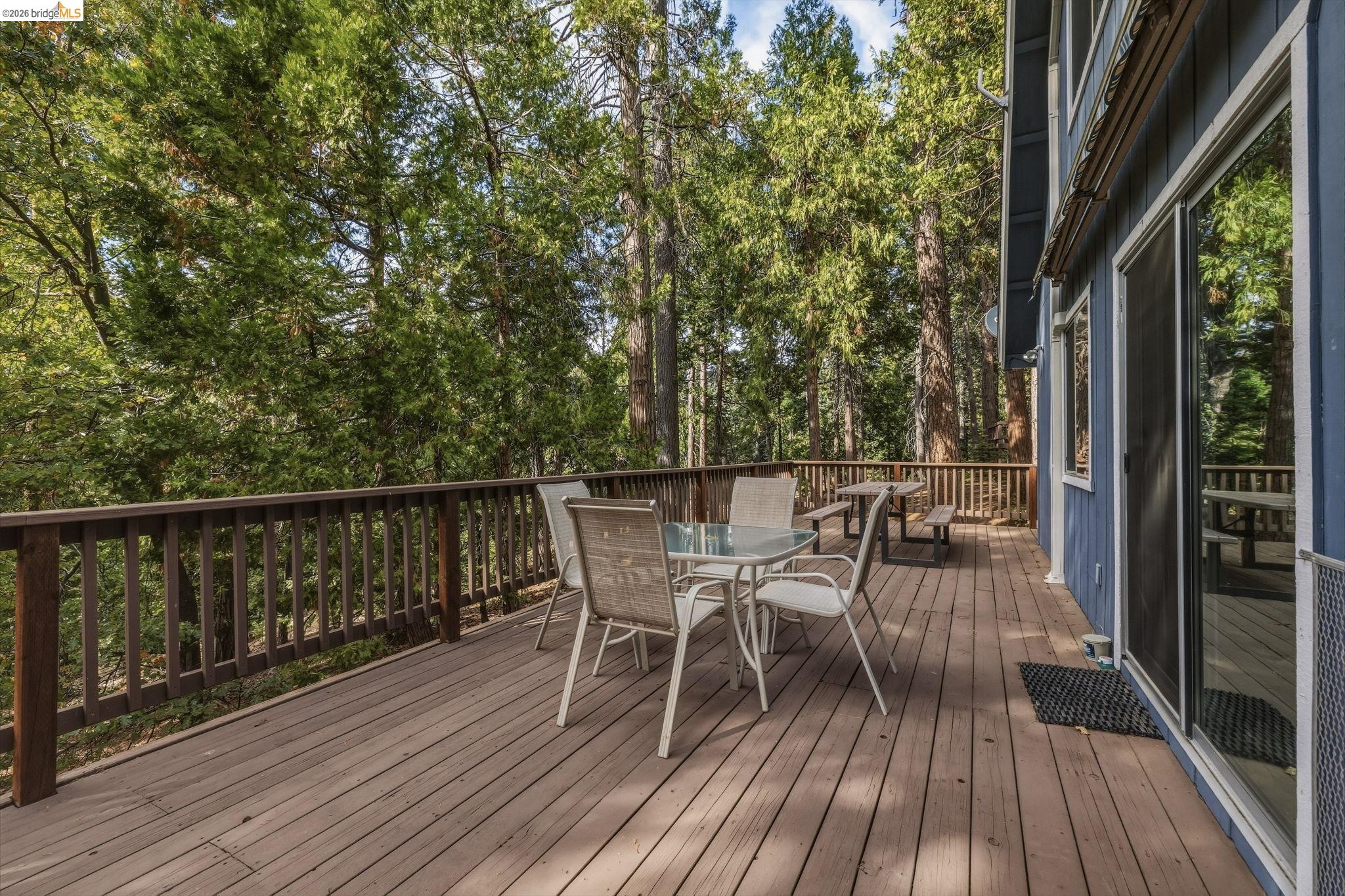 Wooden deck with dining furniture overlooking a dense forest beside a blue-sided home.
