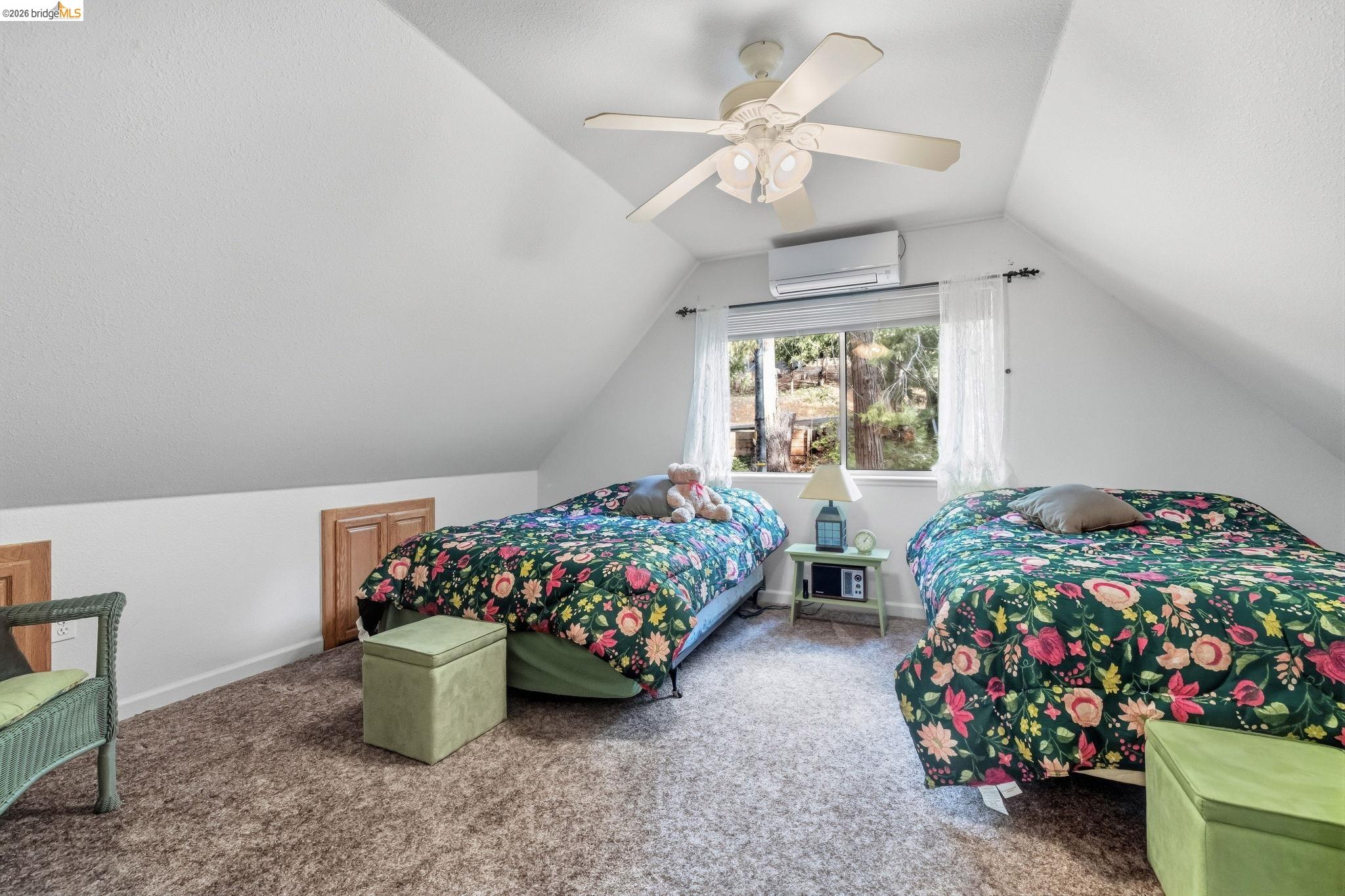 Attic bedroom with two floral-covered twin beds, green ottomans, and a window overlooking trees.