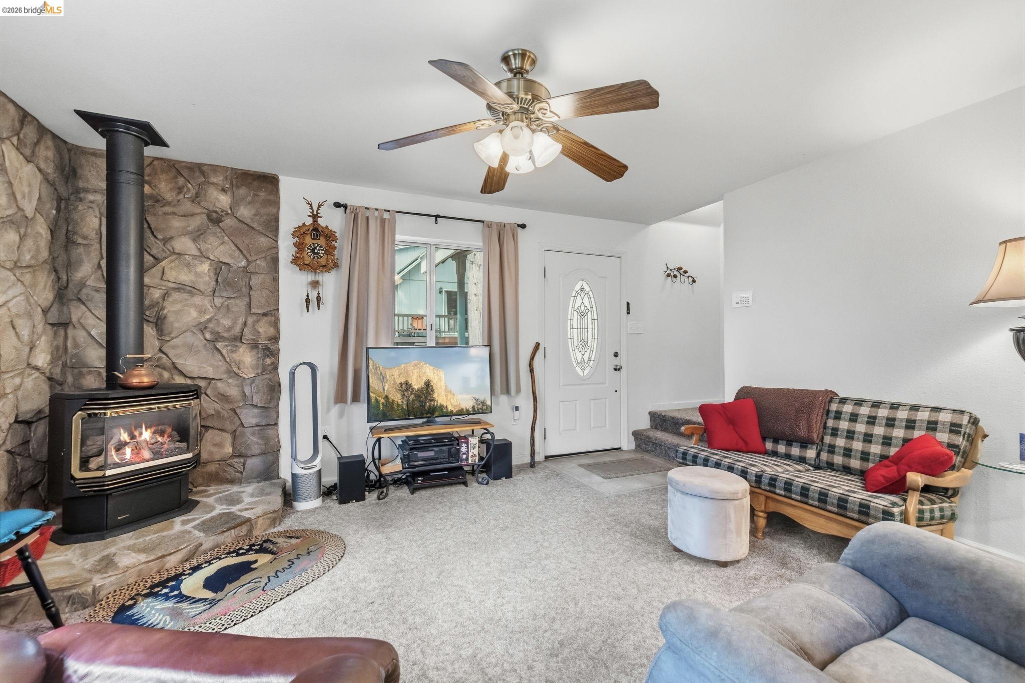 Furnished living room with a stone fireplace and wood-burning stove, plaid sofa, and ceiling fan.