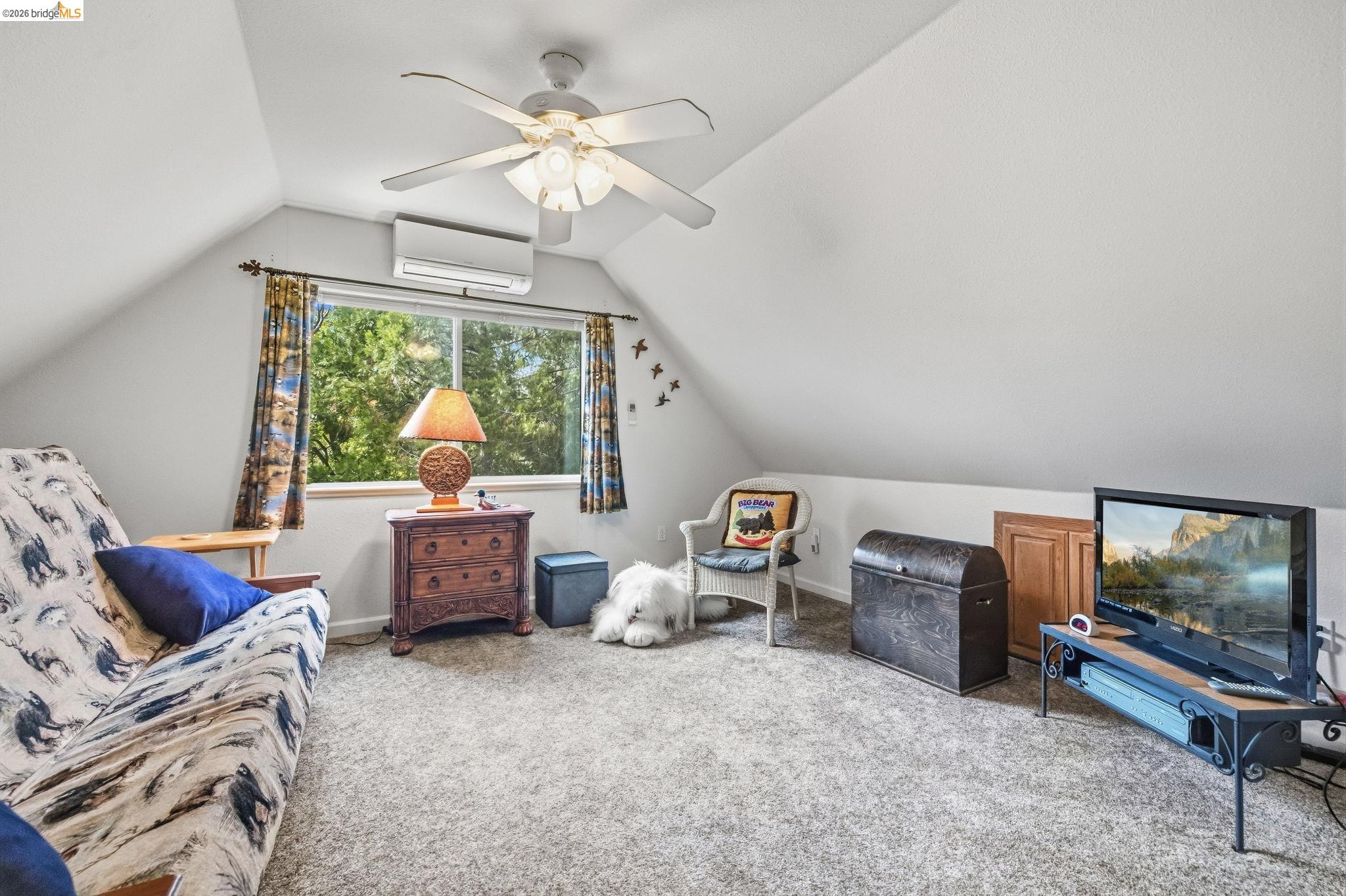 Furnished sitting room with vaulted ceiling, leafy window view, patterned sofa, and television.