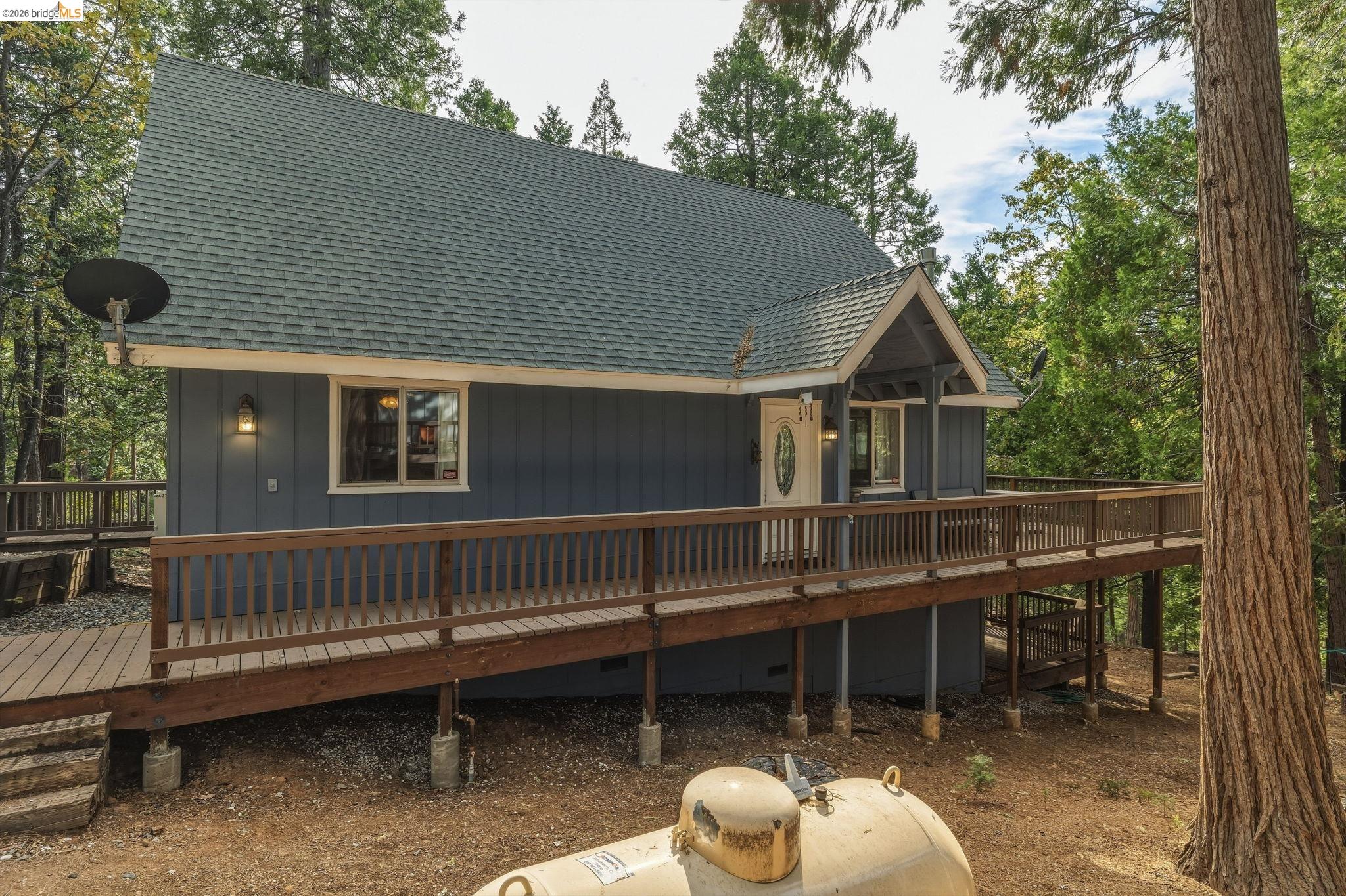 Dark blue-gray house with a steep roof and elevated wooden deck among tall trees.