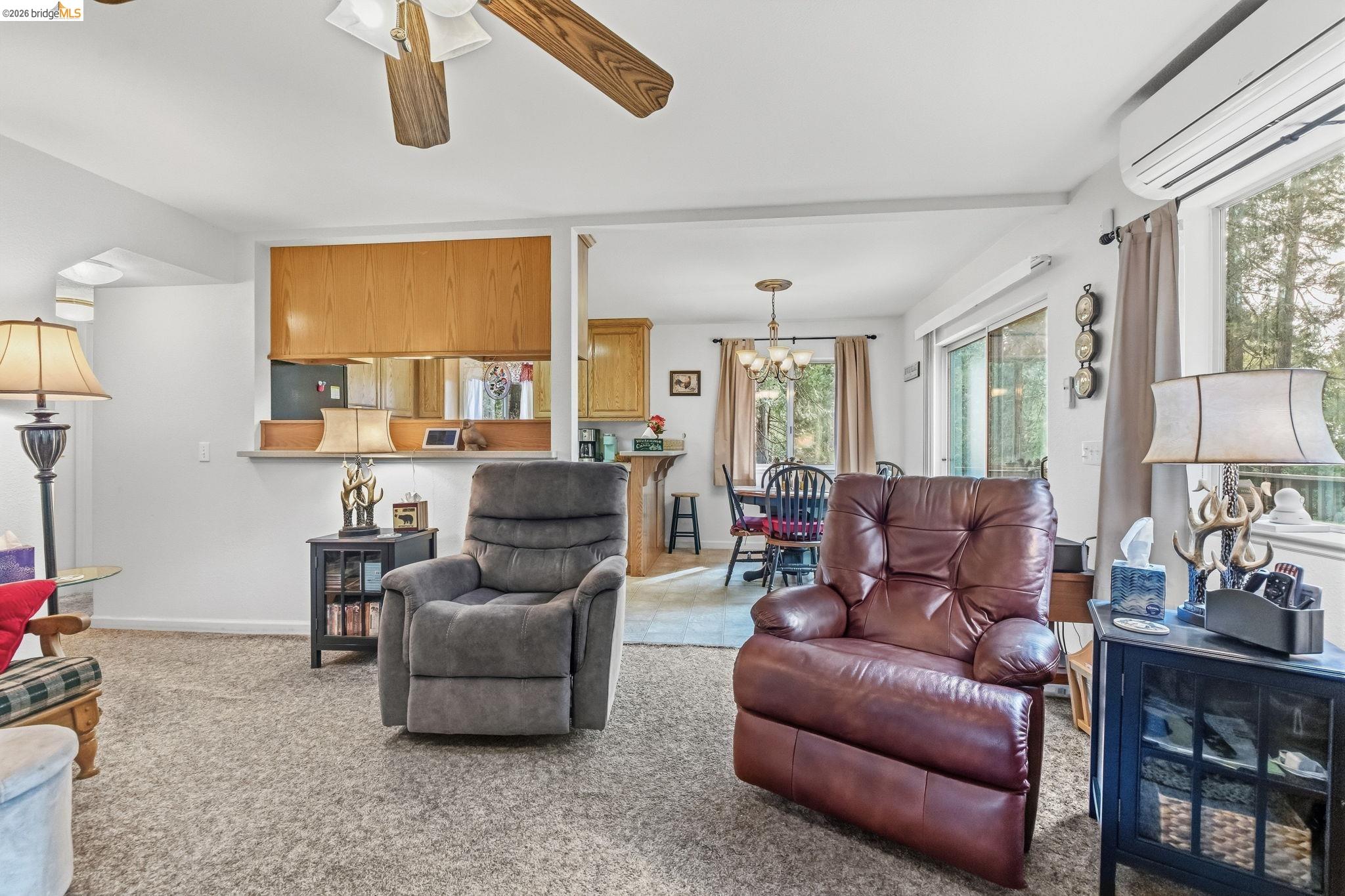 Furnished living room with two recliners, wood kitchen cabinetry, and large windows overlooking trees.