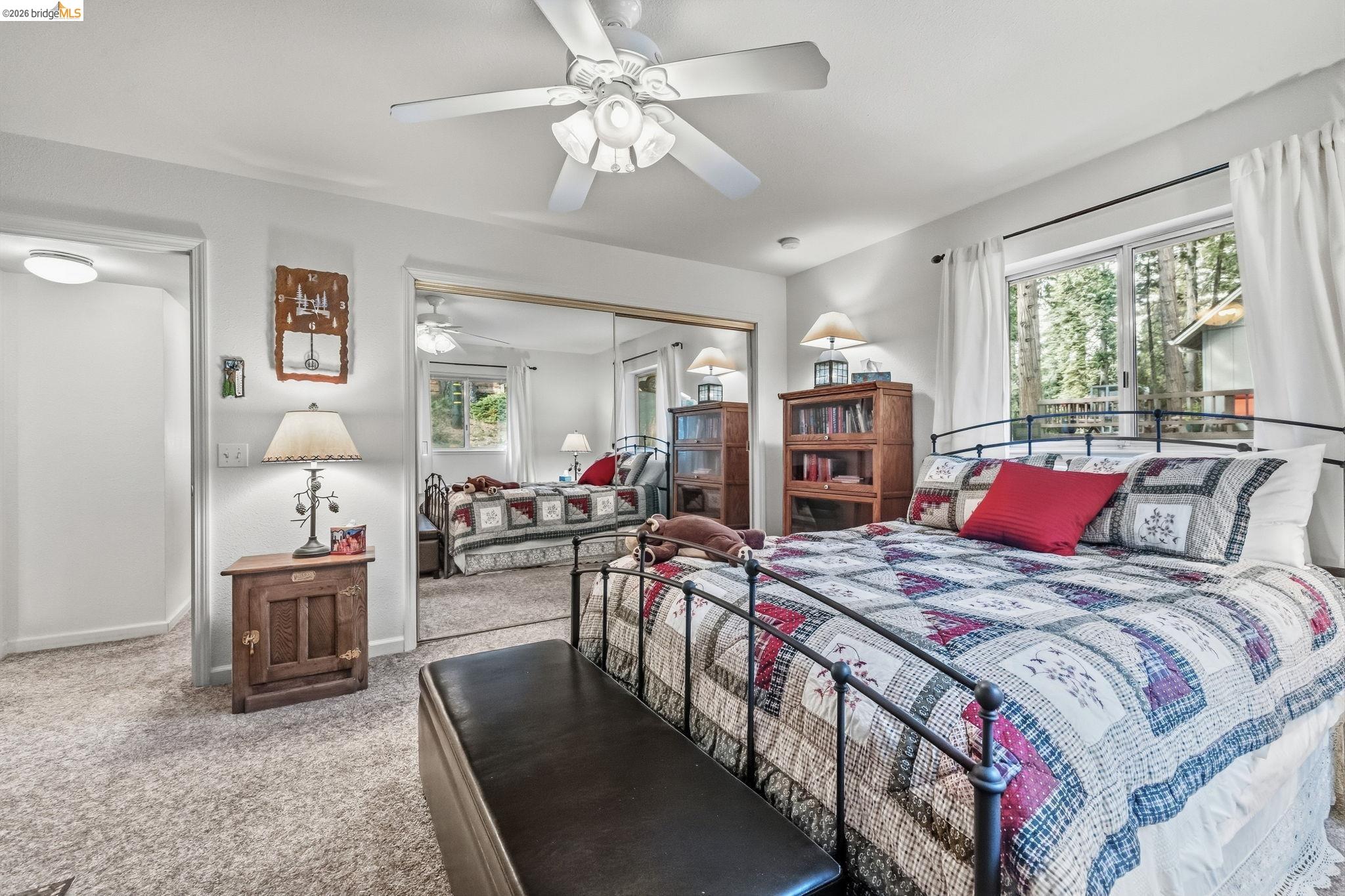Bedroom with a patchwork quilt, mirrored closet doors, and windows overlooking trees.