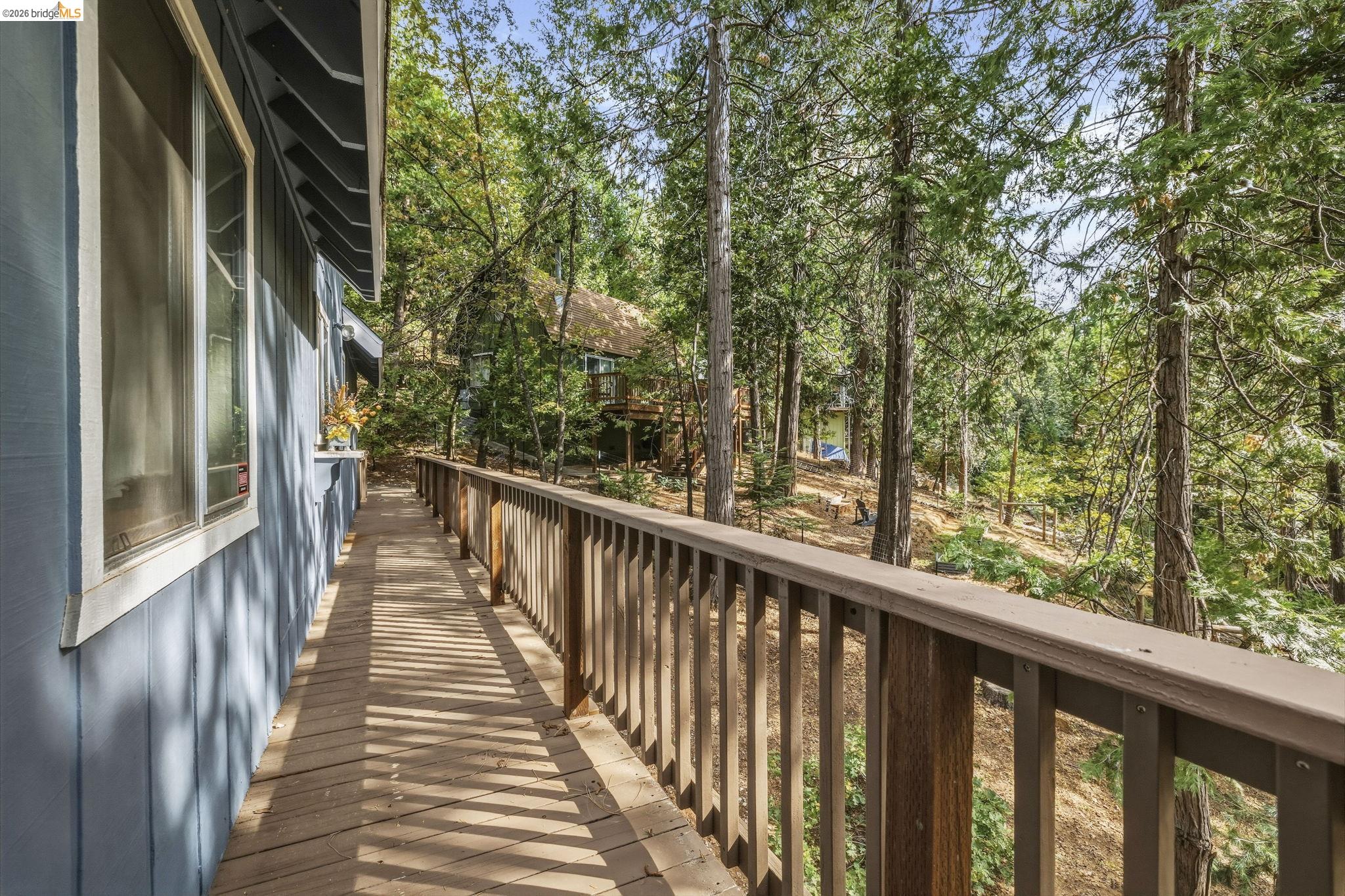 Wooden deck beside a blue-sided home, overlooking a wooded slope and neighboring cabin.