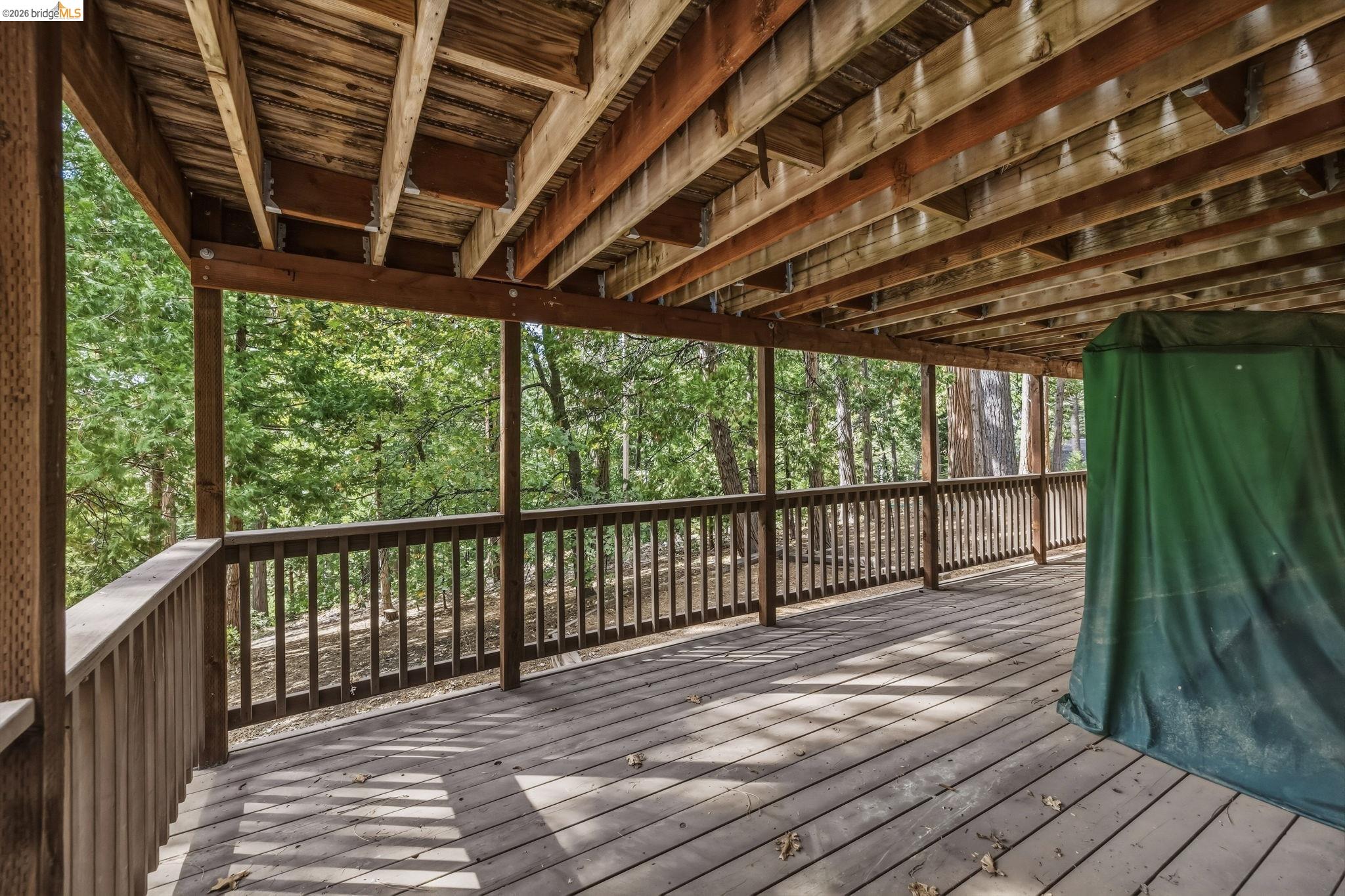 Covered wooden deck with exposed beams and railings overlooking trees, with a green tarp-covered object at one end.