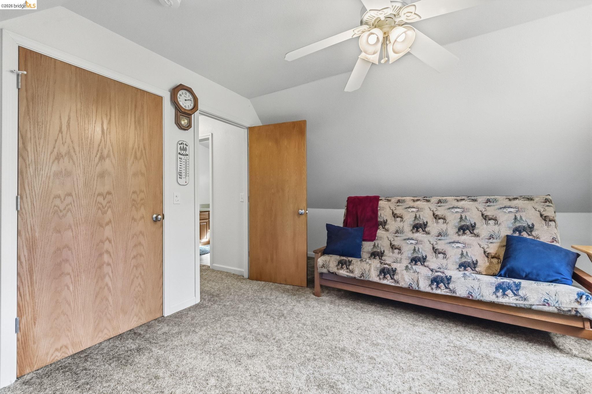 Carpeted room with a patterned sofa bed, sloped ceiling, wooden doors, and ceiling fan.
