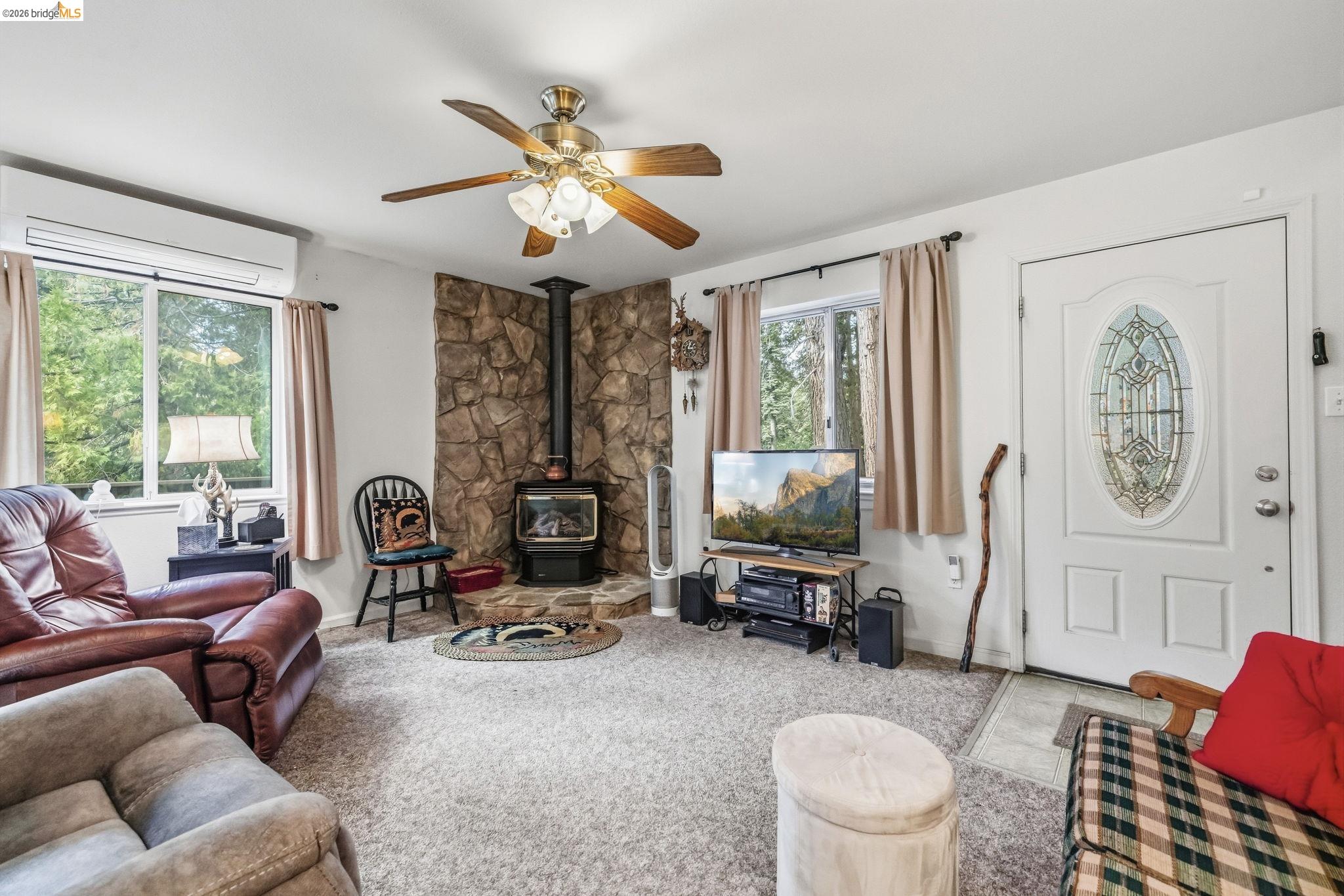 Furnished living room with a stone fireplace and wood-burning stove, ceiling fan, and windows overlooking trees.