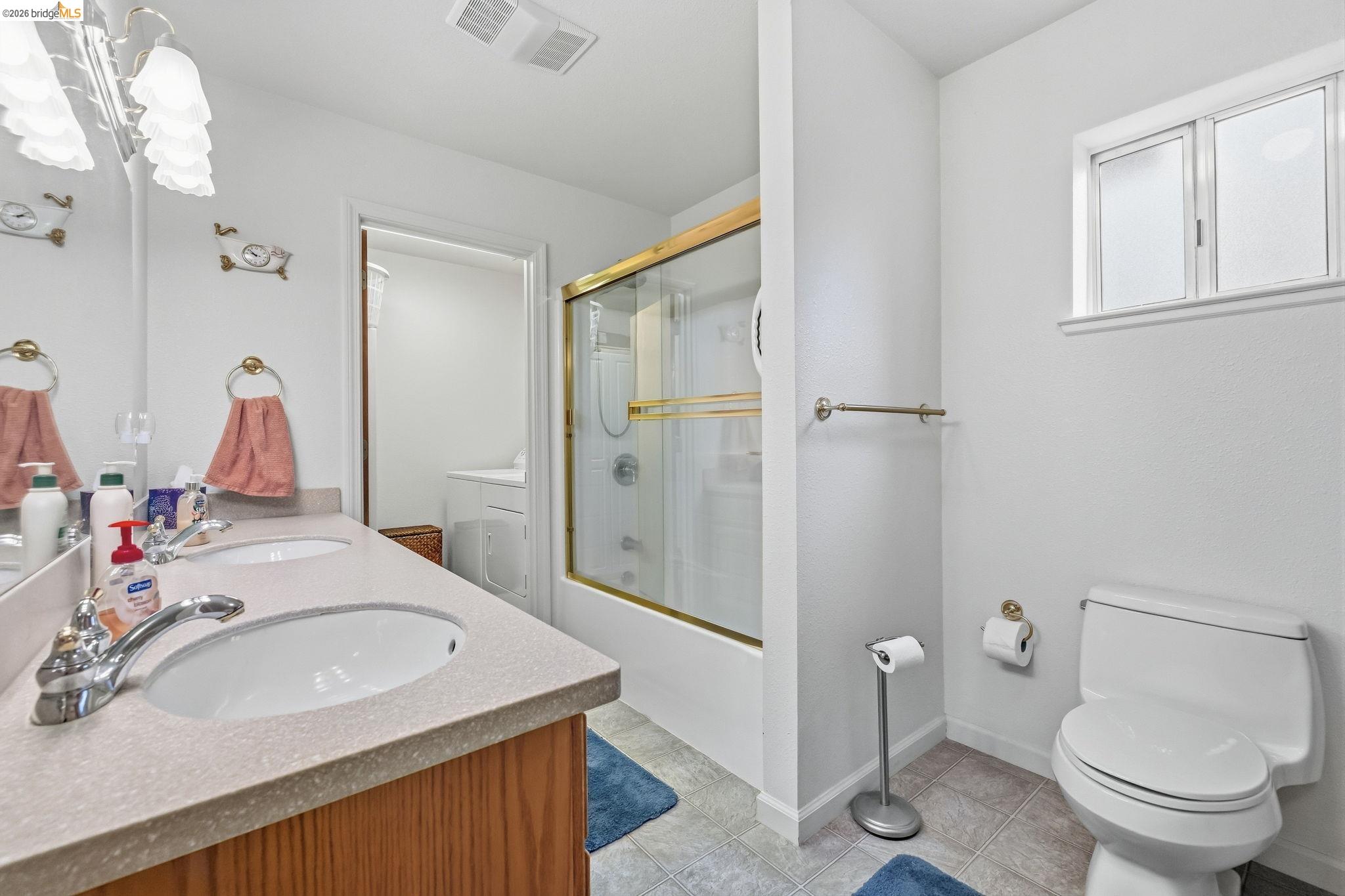 Bathroom with a double-sink vanity, gold-framed glass shower, and toilet beneath a frosted window.