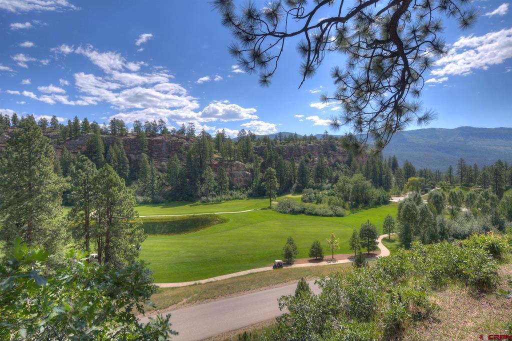 Golf course with a pond and winding paths, framed by pine trees and distant mountains.