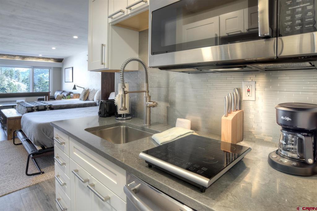Compact condo kitchen with gray counters, stainless appliances, and two beds in the adjoining room.