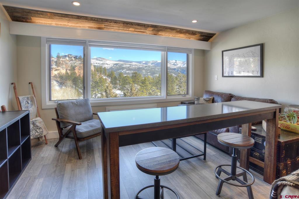 Furnished dining area with a large window overlooking snowy mountains and pine-covered hills.