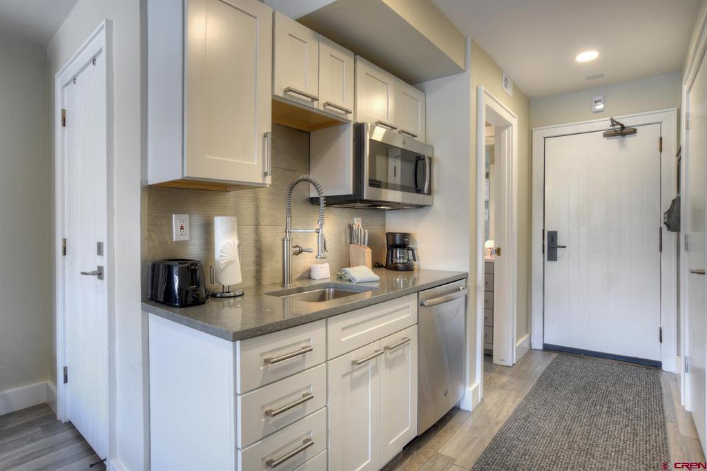 Compact kitchenette with white cabinets, gray counters, stainless-steel appliances, and the condo entry door.
