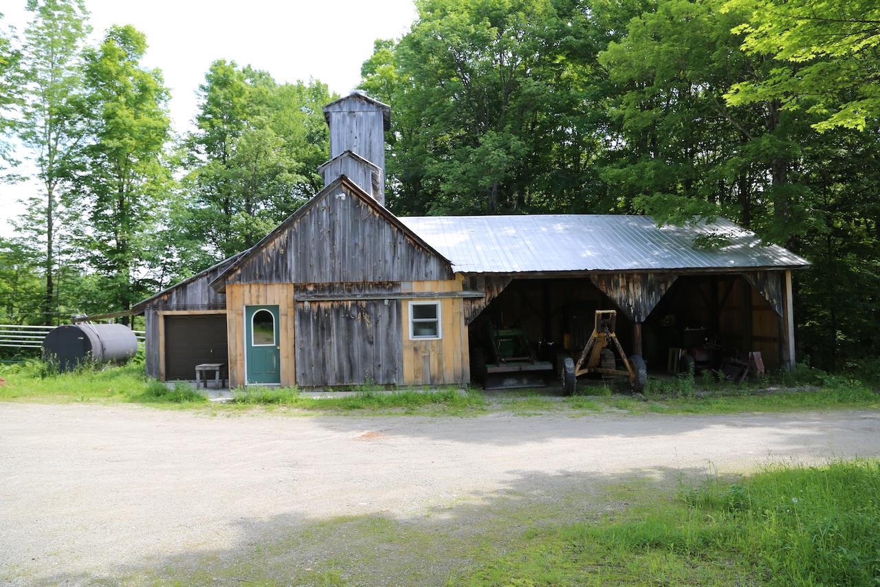 Weathered wooden barn with a metal roof, green door, and open equipment bays among trees.